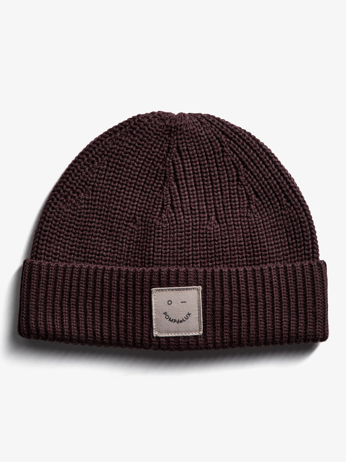 A knitted beanie resting flat on a white surface features a visible patch with a smiley face and "POMPdeLUX" text on the folded brim.