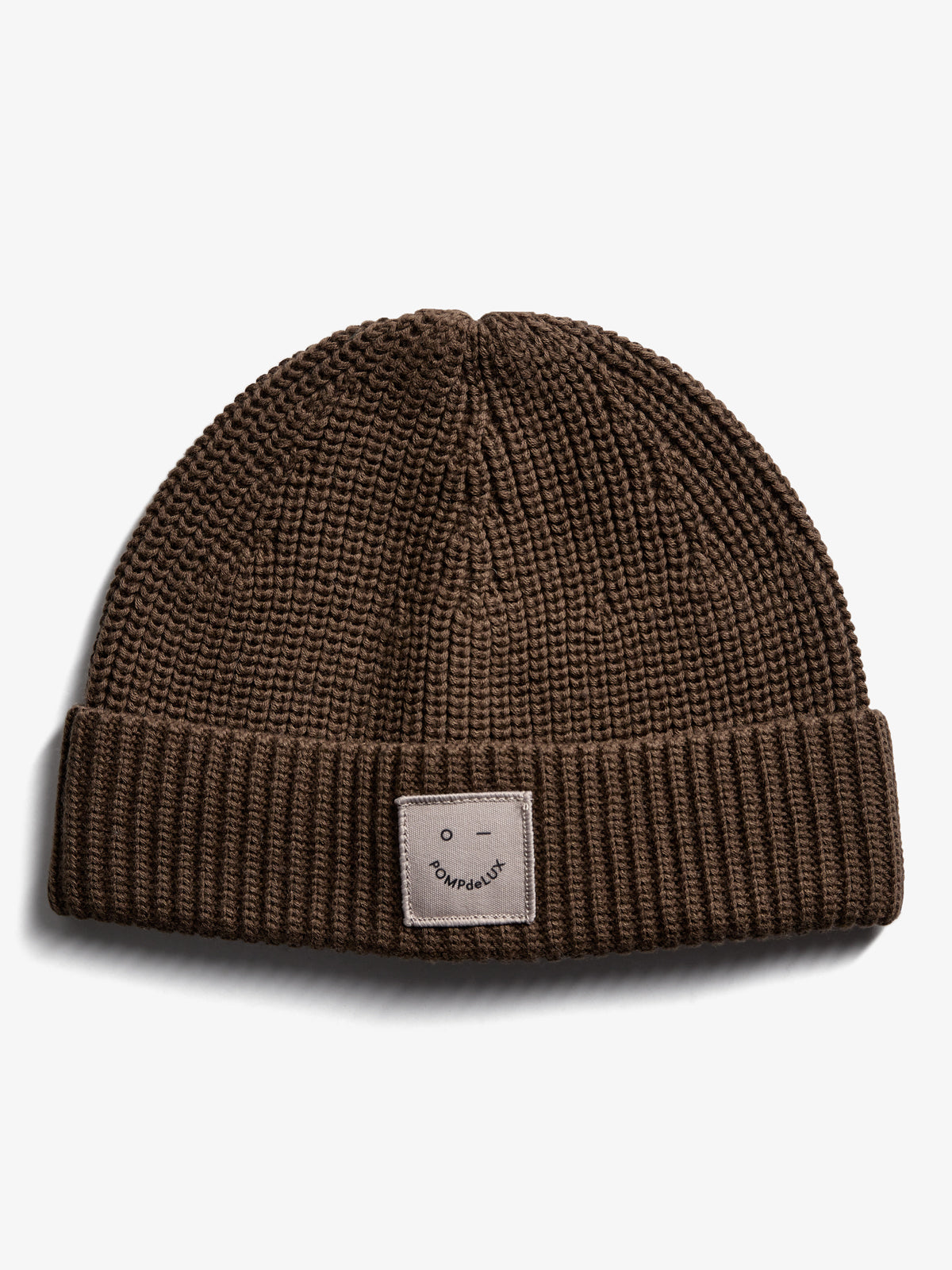 Brown knitted beanie lies flat, featuring a folded brim and a small patch with a minimalist face and text: "POMPdeLUX." Set against a plain white background.
