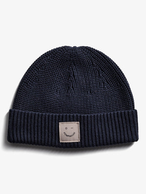 A navy blue knitted beanie lies flat against a white background. The cuff features a sewn patch with a simple face illustration and the text "POMPdeLUX."