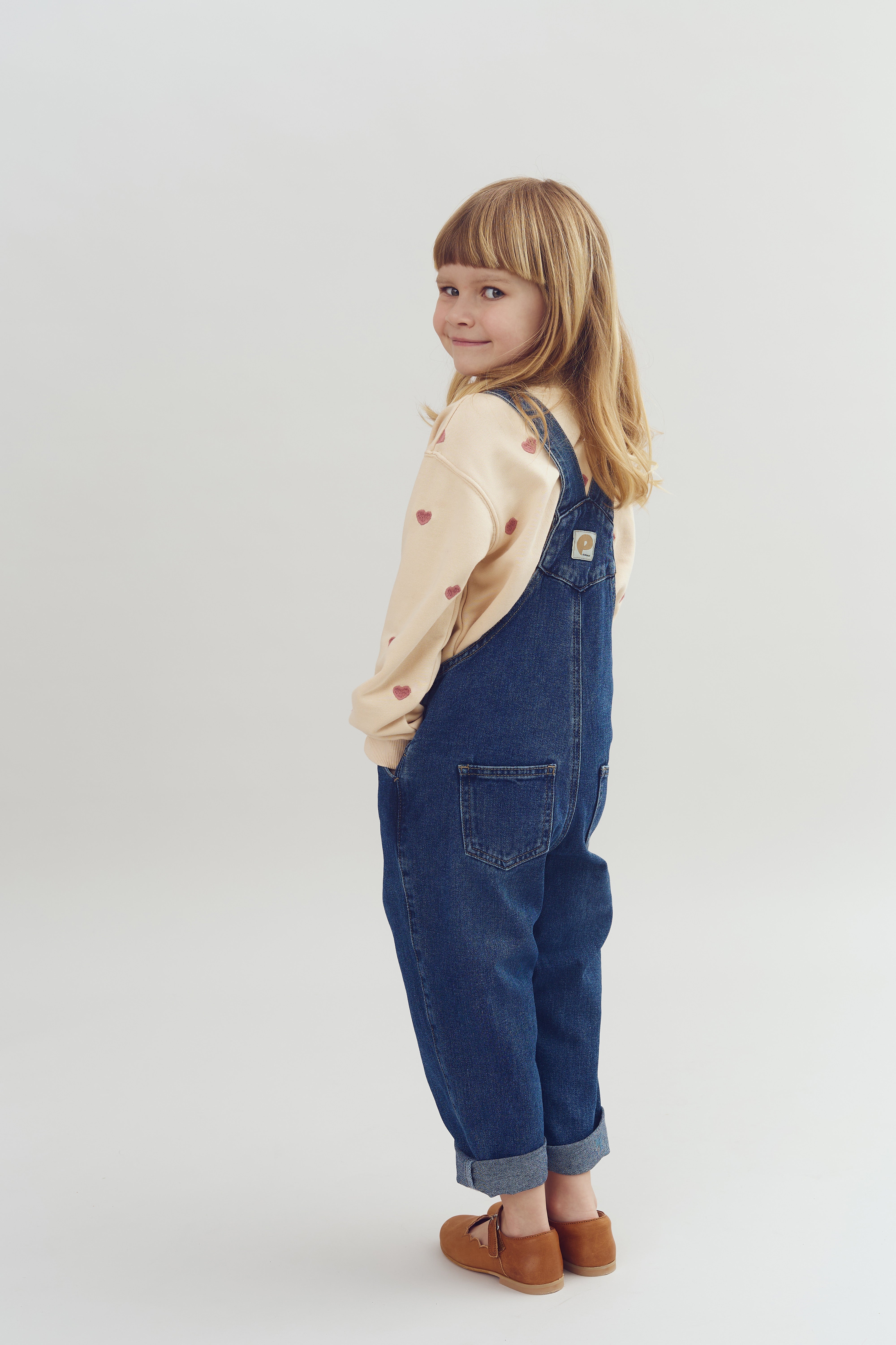 Kind zeigt ein sanftes Lächeln, während es in einer Jeans-Latzhose und einem beige-farbenen Pullover mit Herzen posiert. Der Hintergrund ist neutral und hell.