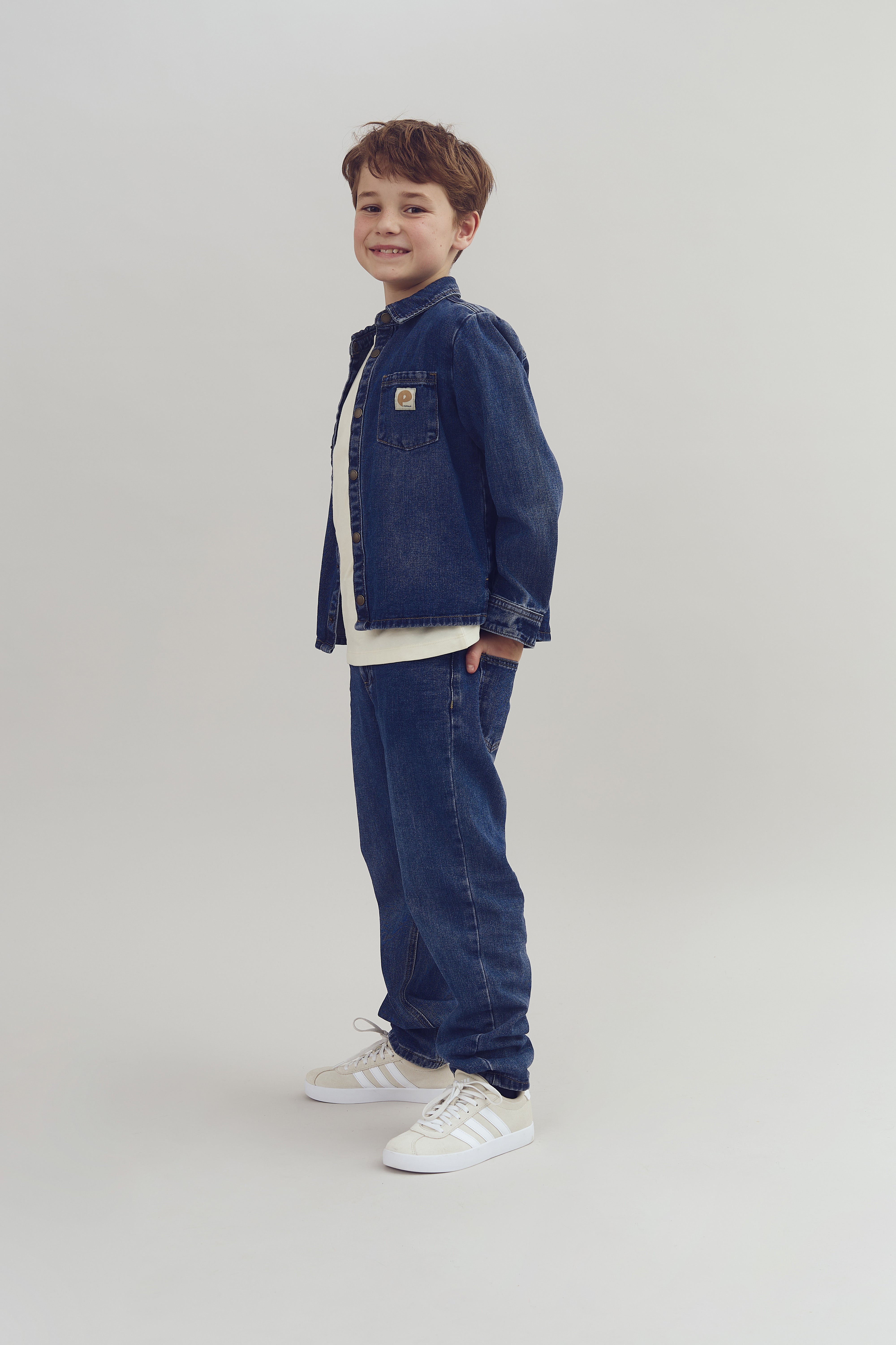 A child wearing a denim jacket and jeans smiles while standing sideways in a bright, minimalist studio. They're wearing white sneakers and a simple shirt underneath the jacket.