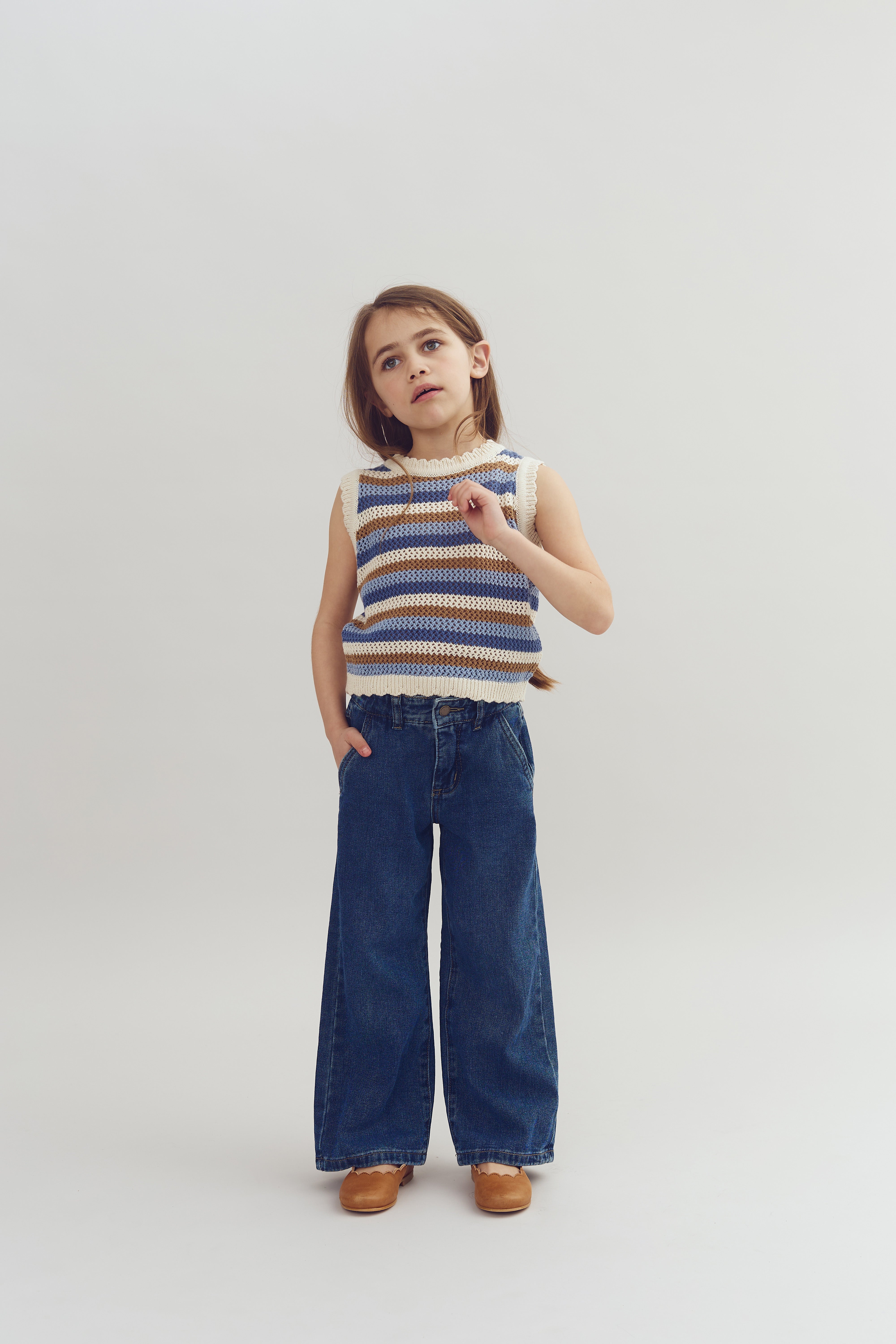 Ein Kind steht und schaut nachdenklich, trägt ein gestreiftes Oberteil und weit geschnittene Jeans. Die rechte Hand steckt in der Hosentasche, der Hintergrund ist schlicht und neutral.