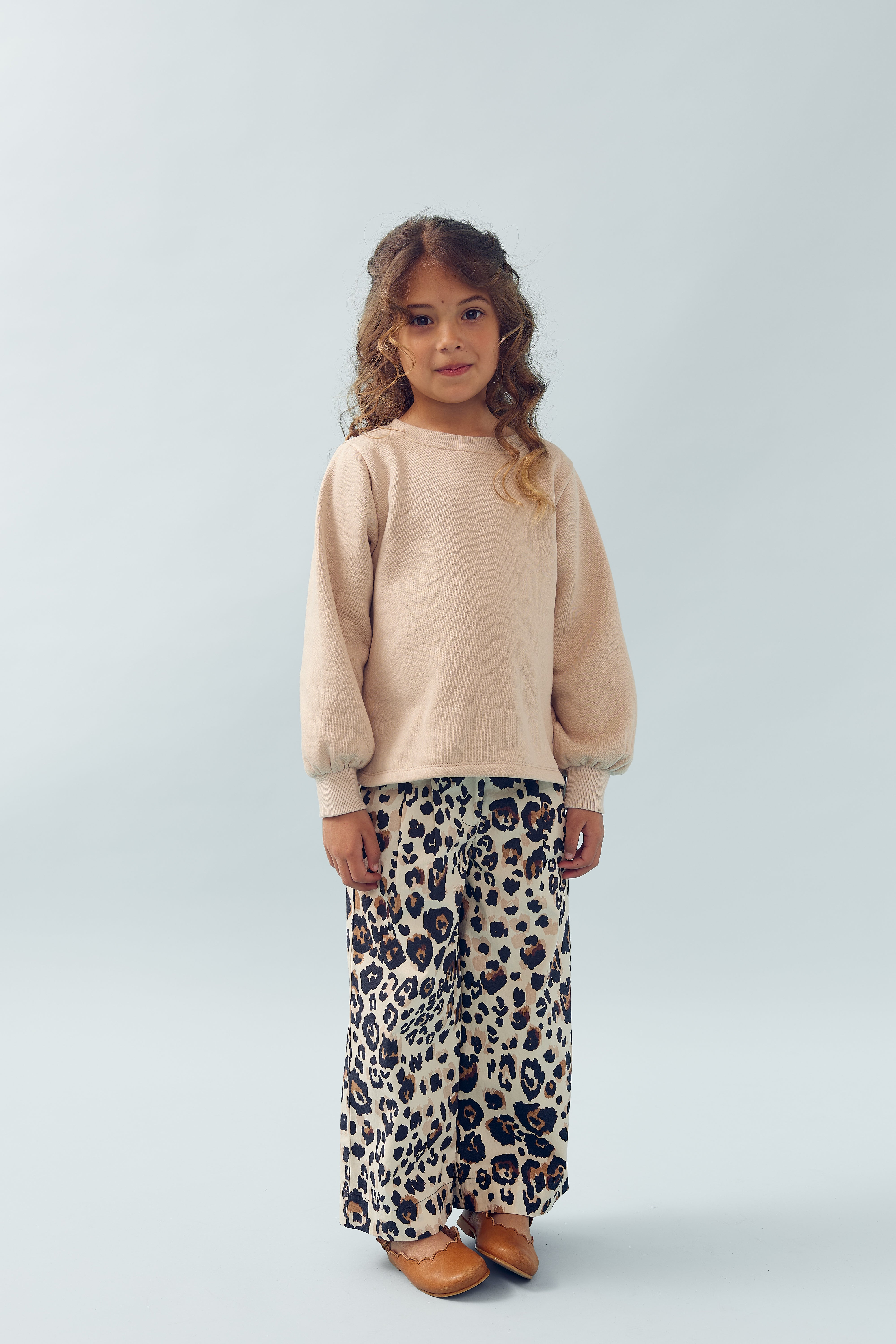 Ein Kind steht ruhig im Raum, trägt einen beigen Pullover und weite Hosen mit Leopardenmuster. Der Hintergrund ist schlicht und einfarbig hell.