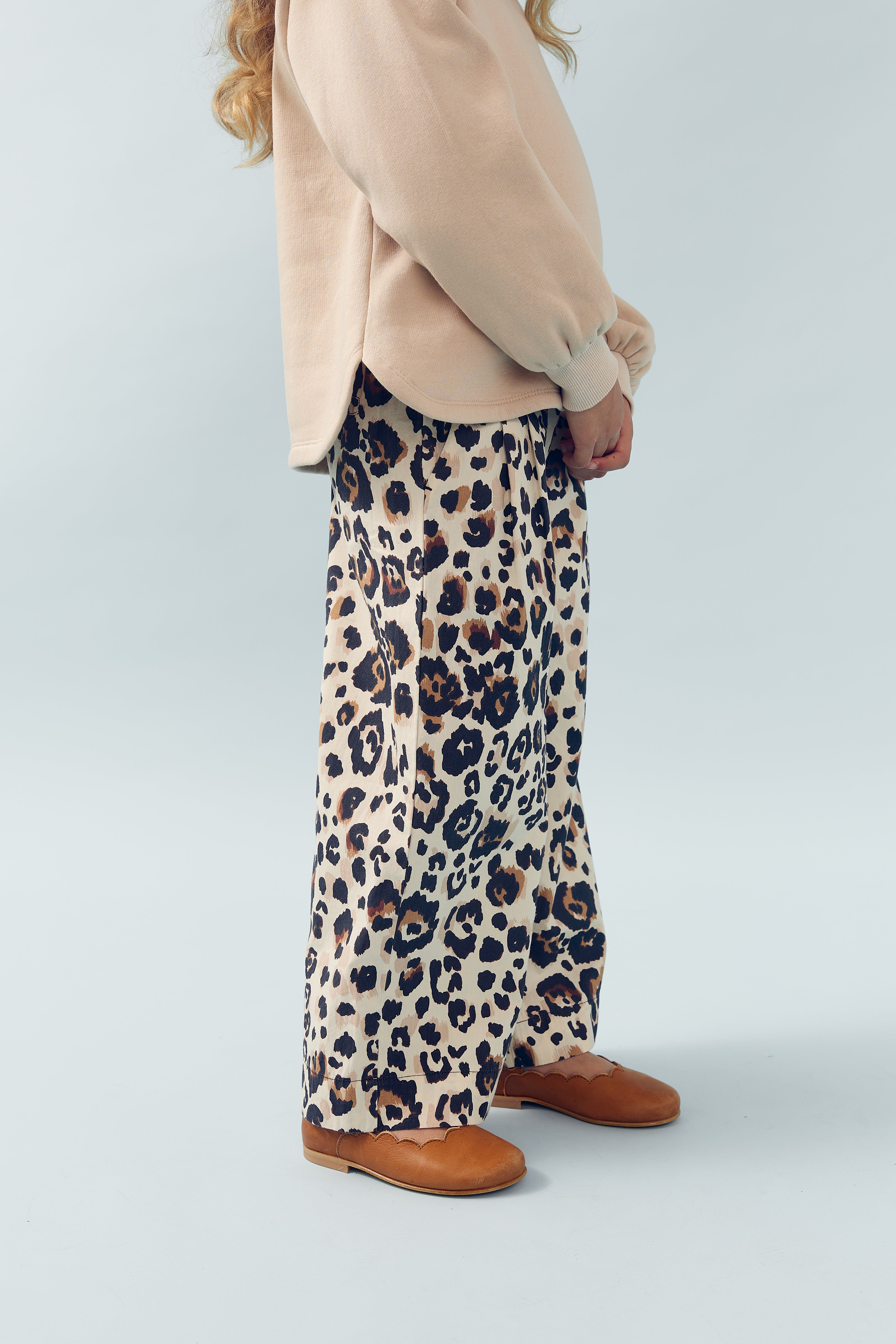 Ein Kind trägt ein beiges Oberteil und weite, leopardengemusterte Hosen mit braunen Schuhen. Es steht aufrecht in einem hellgrauen, minimalistischen Studiohintergrund.