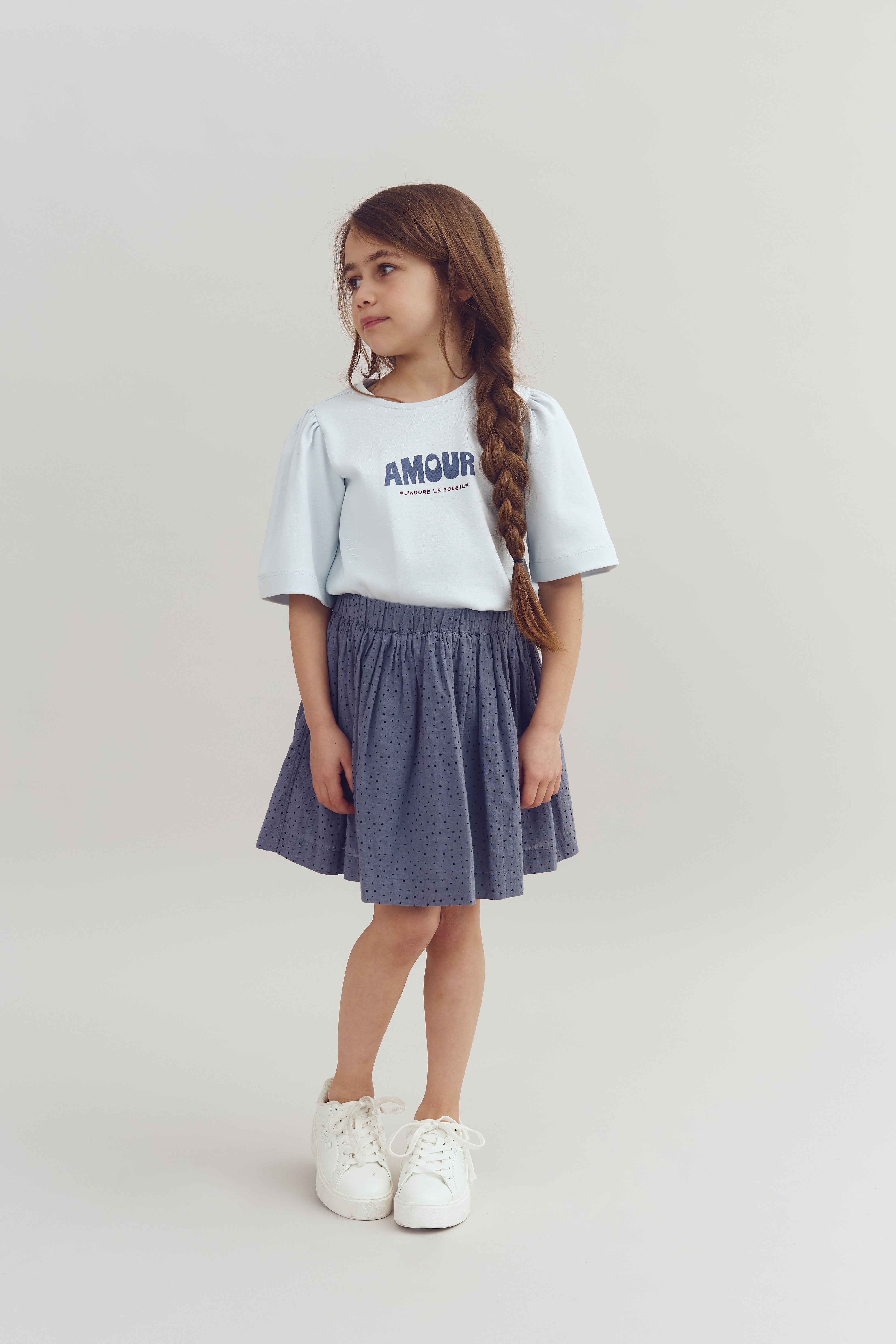 A young girl stands wearing a light blue shirt with "AMOUR J'ADORE LE MODELE" printed on it, paired with a gray skirt and white sneakers, against a plain background.