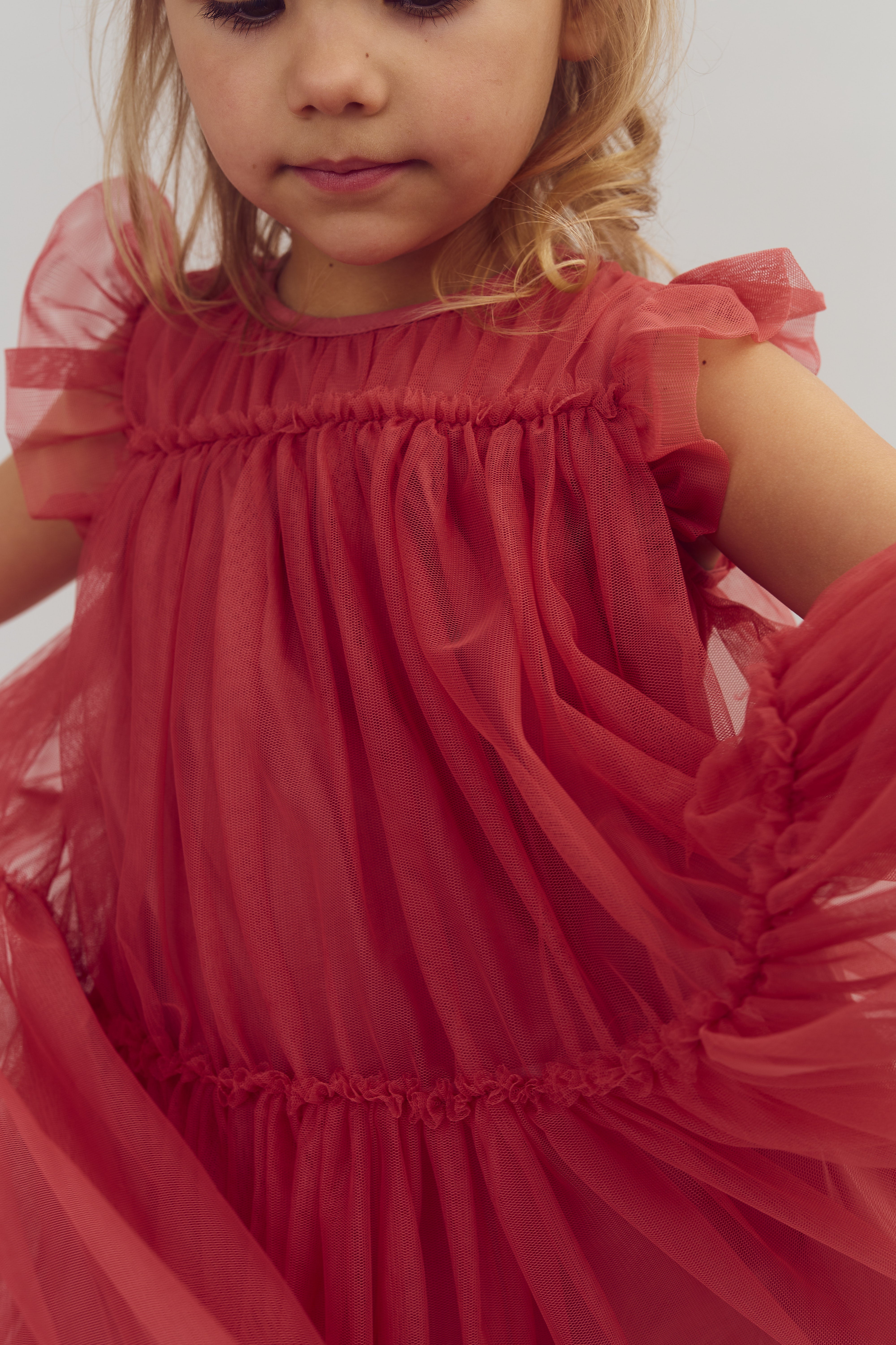 A child with light hair wears a frilly, sheer red dress, with one arm raised. The background is a light, neutral color.