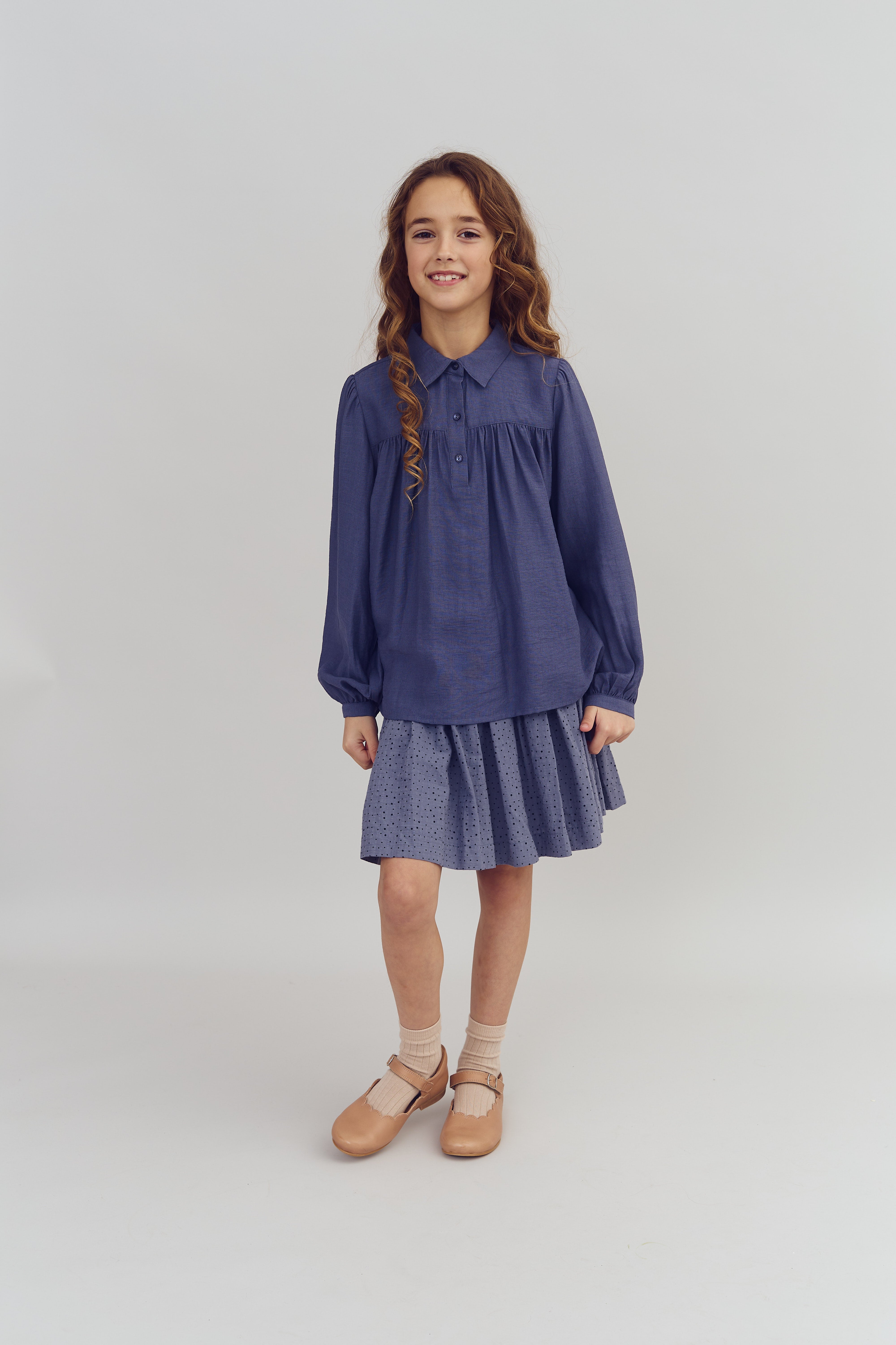 Ein Mädchen steht lächelnd in einem blauen Kleid mit langen Ärmeln und hellbraunen Schuhen, umgeben von einem neutralen Hintergrund.