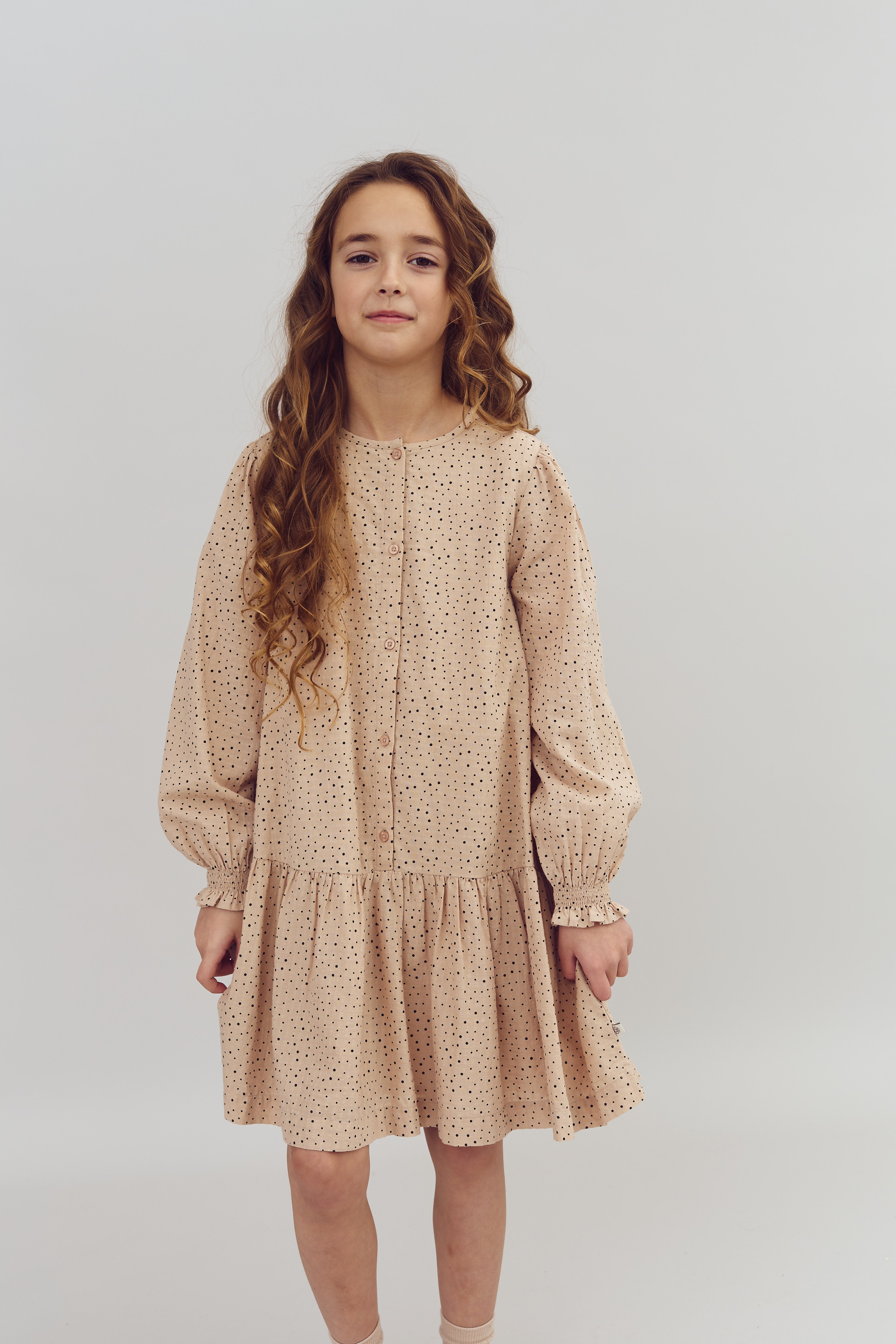 A young girl stands with long, curly hair, wearing a light pink, polka-dotted dress in a plain white studio setting.