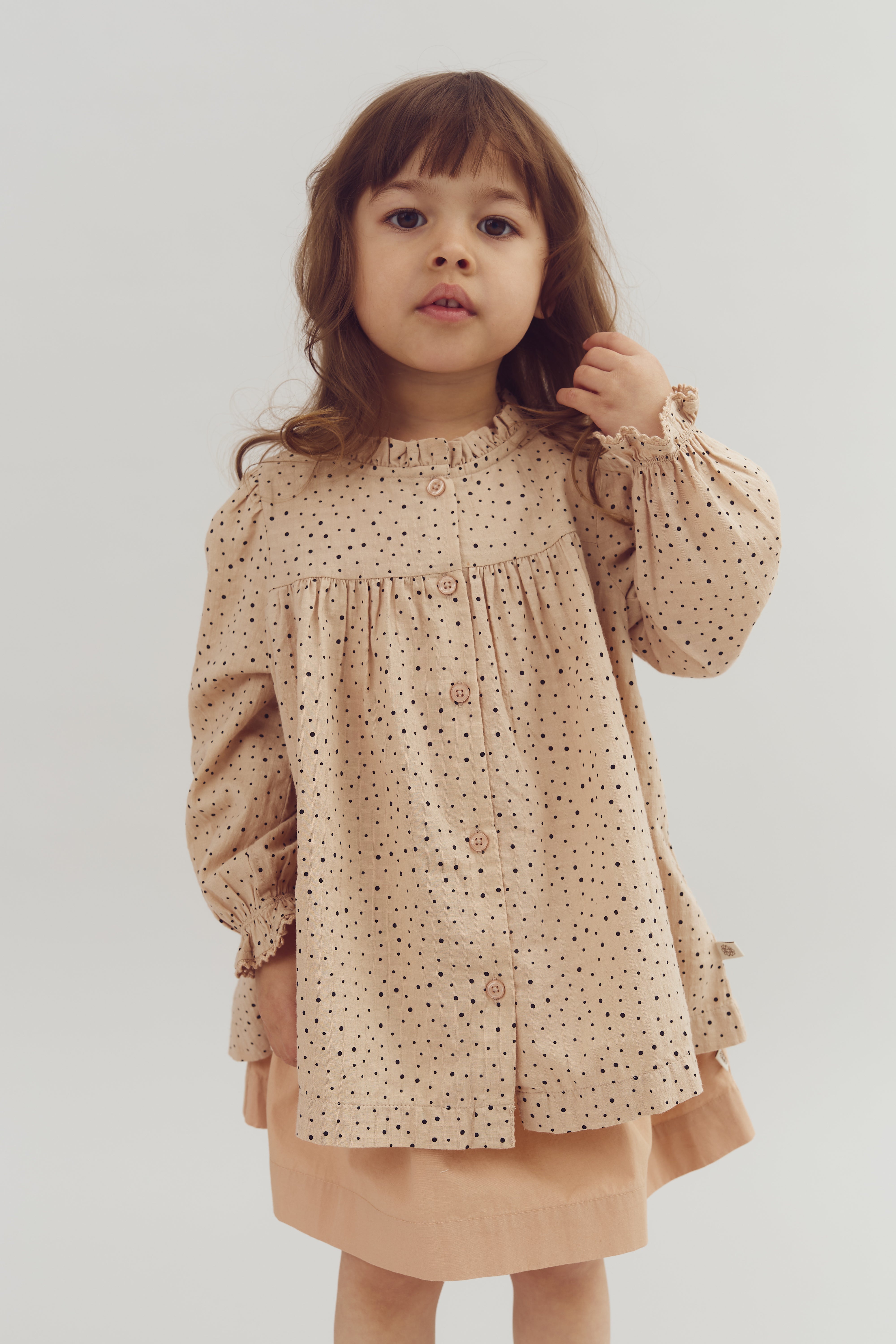 Child wearing a beige, polka-dotted dress touches their hair, standing against a plain white background.