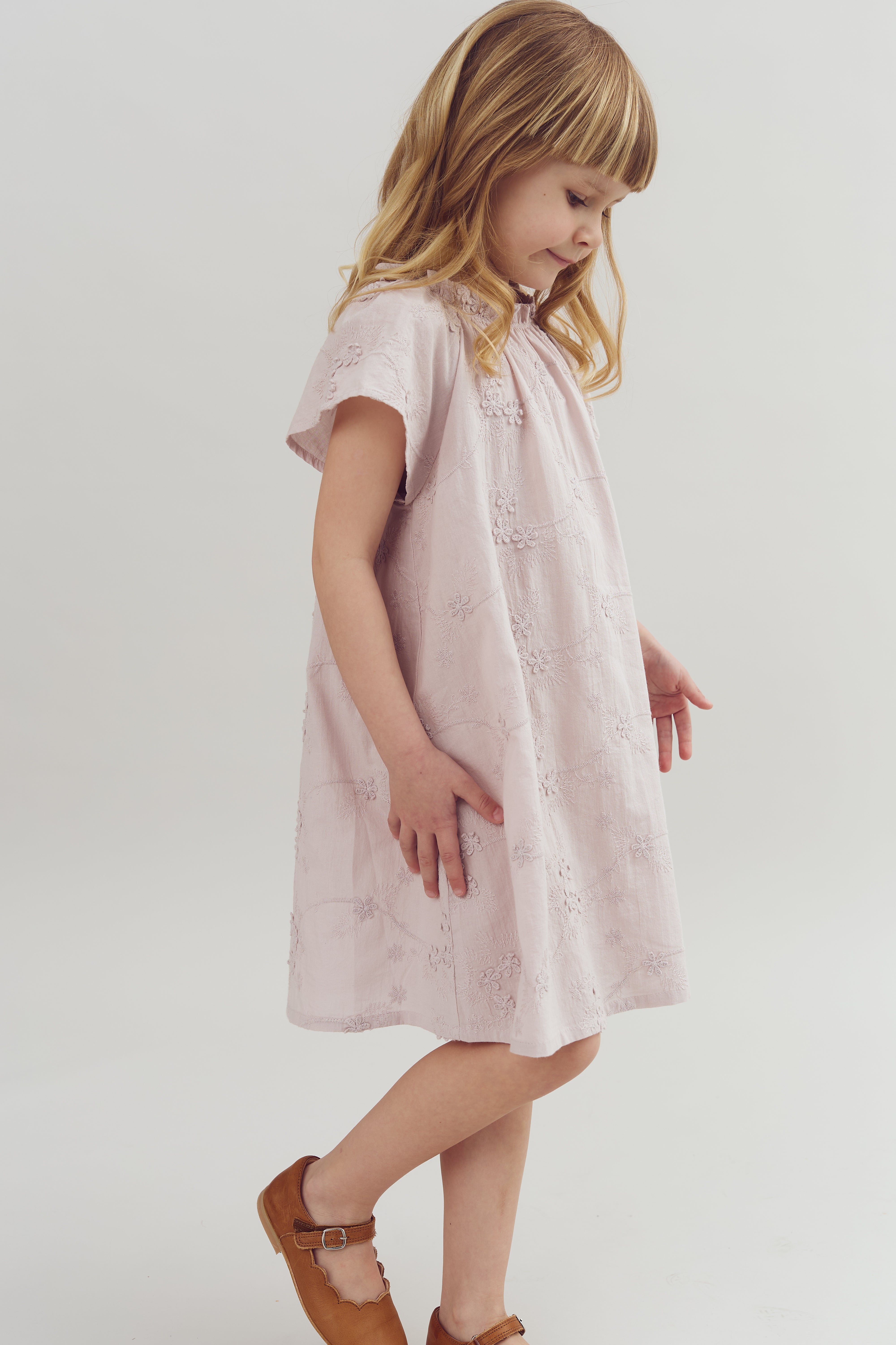 A young child in a light pink dress with floral patterns walks gracefully, wearing brown shoes, in a plain white studio setting.