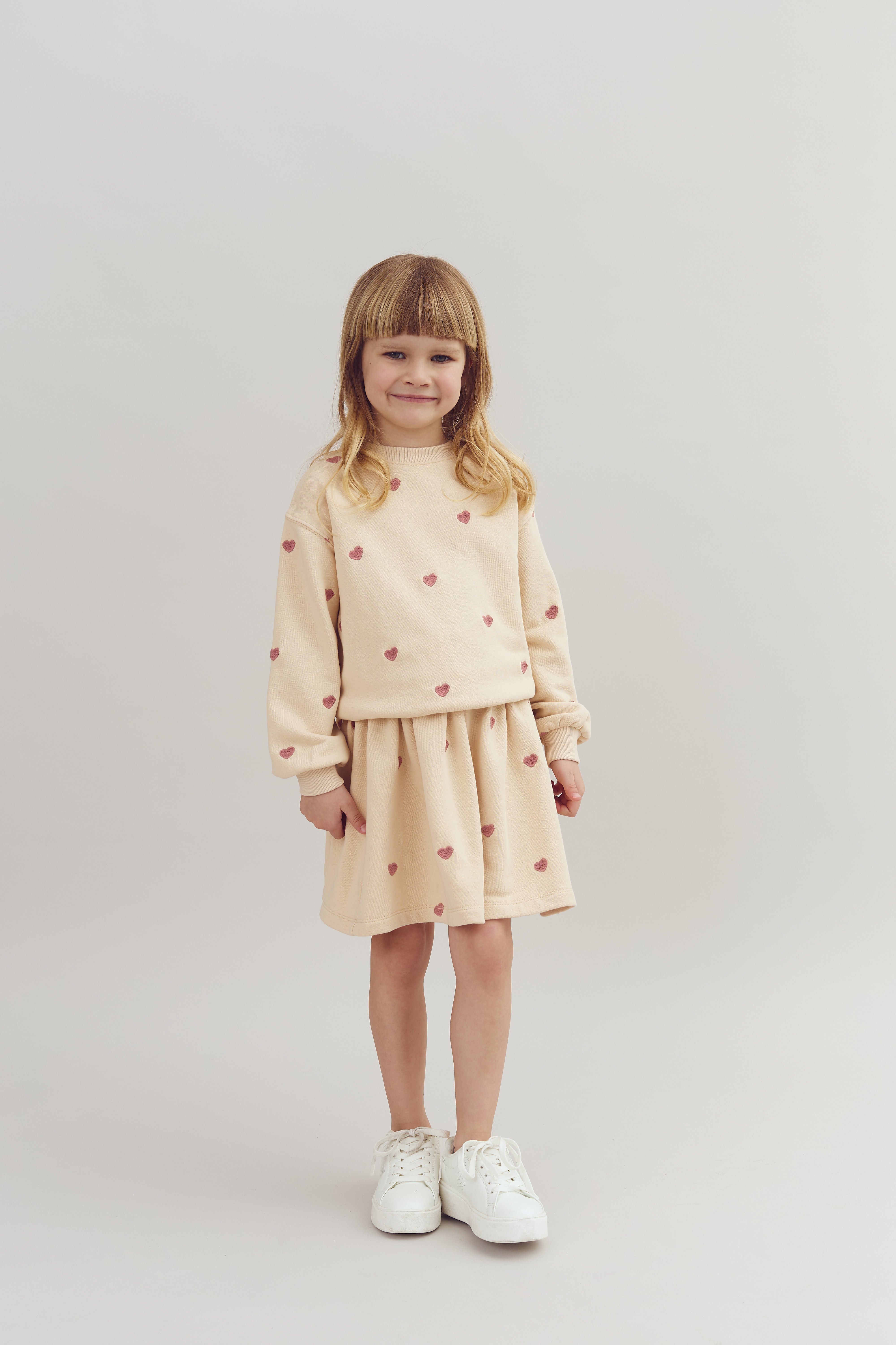 A young child stands smiling, wearing a beige outfit with pink hearts and white sneakers, against a plain light background.
