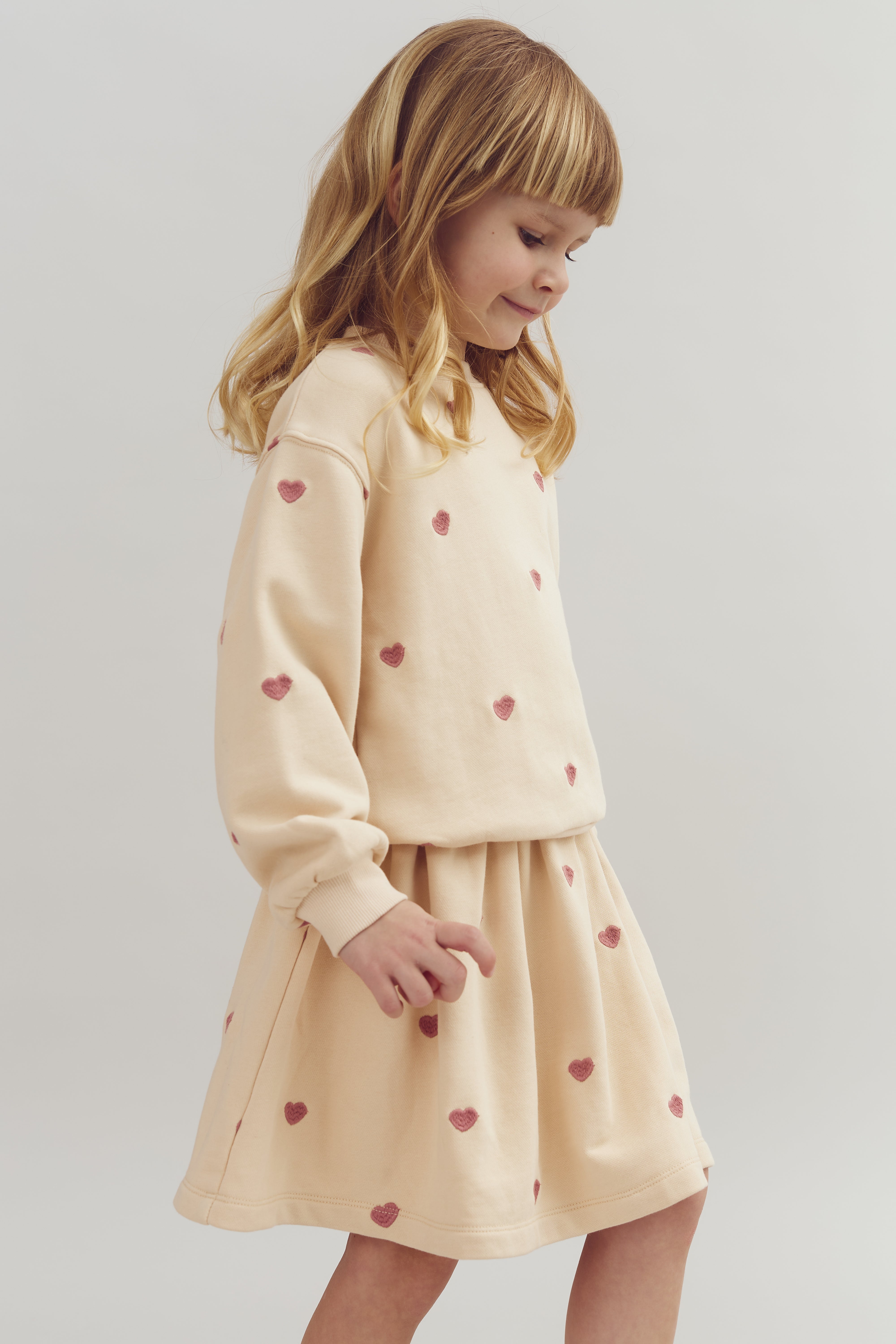 A child wearing a cream-colored outfit with red heart patterns stands in a side profile, against a plain light gray background.