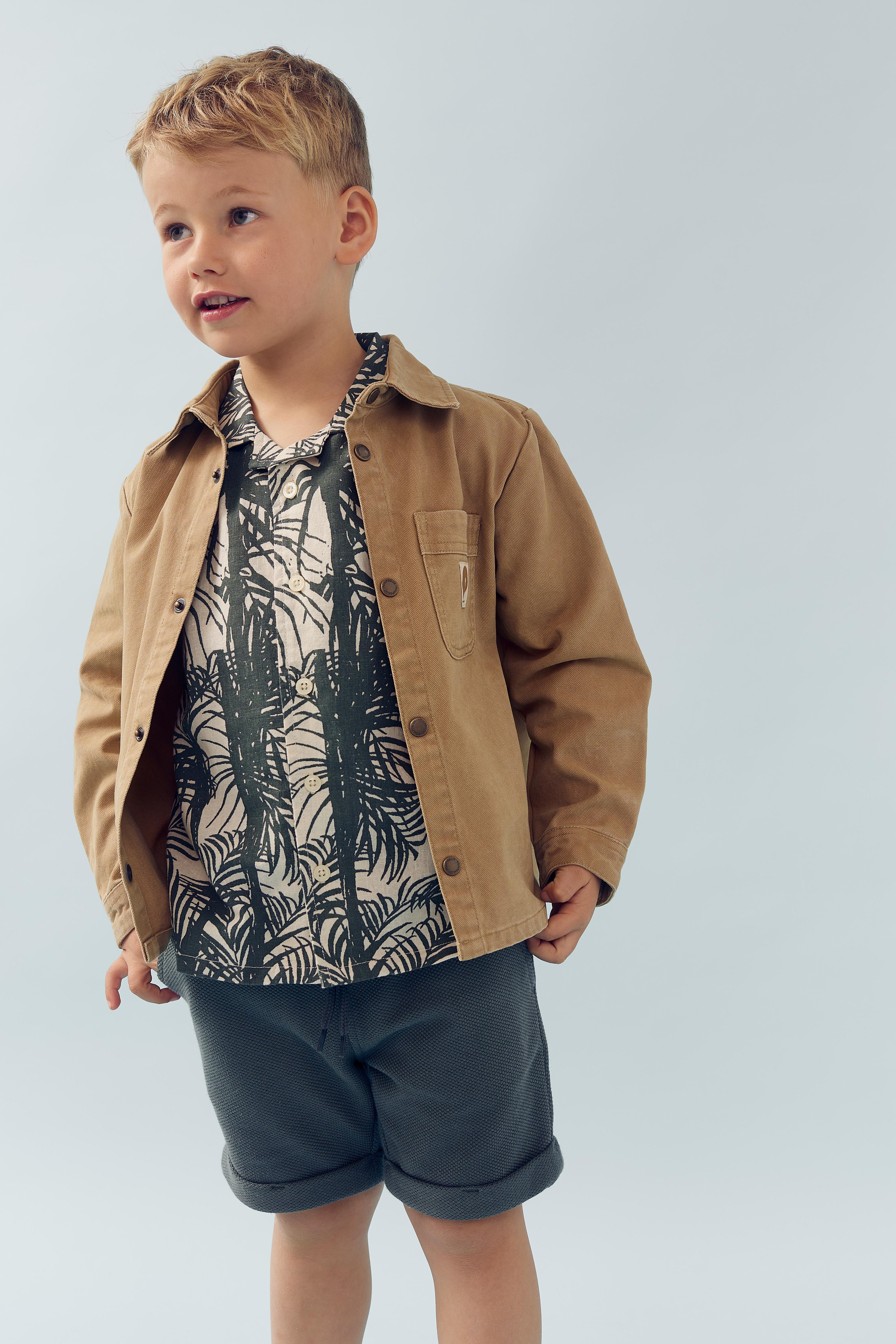 A young boy wears a brown jacket, patterned shirt, and gray shorts, standing against a plain light gray background. He looks to the side with a slight smile.