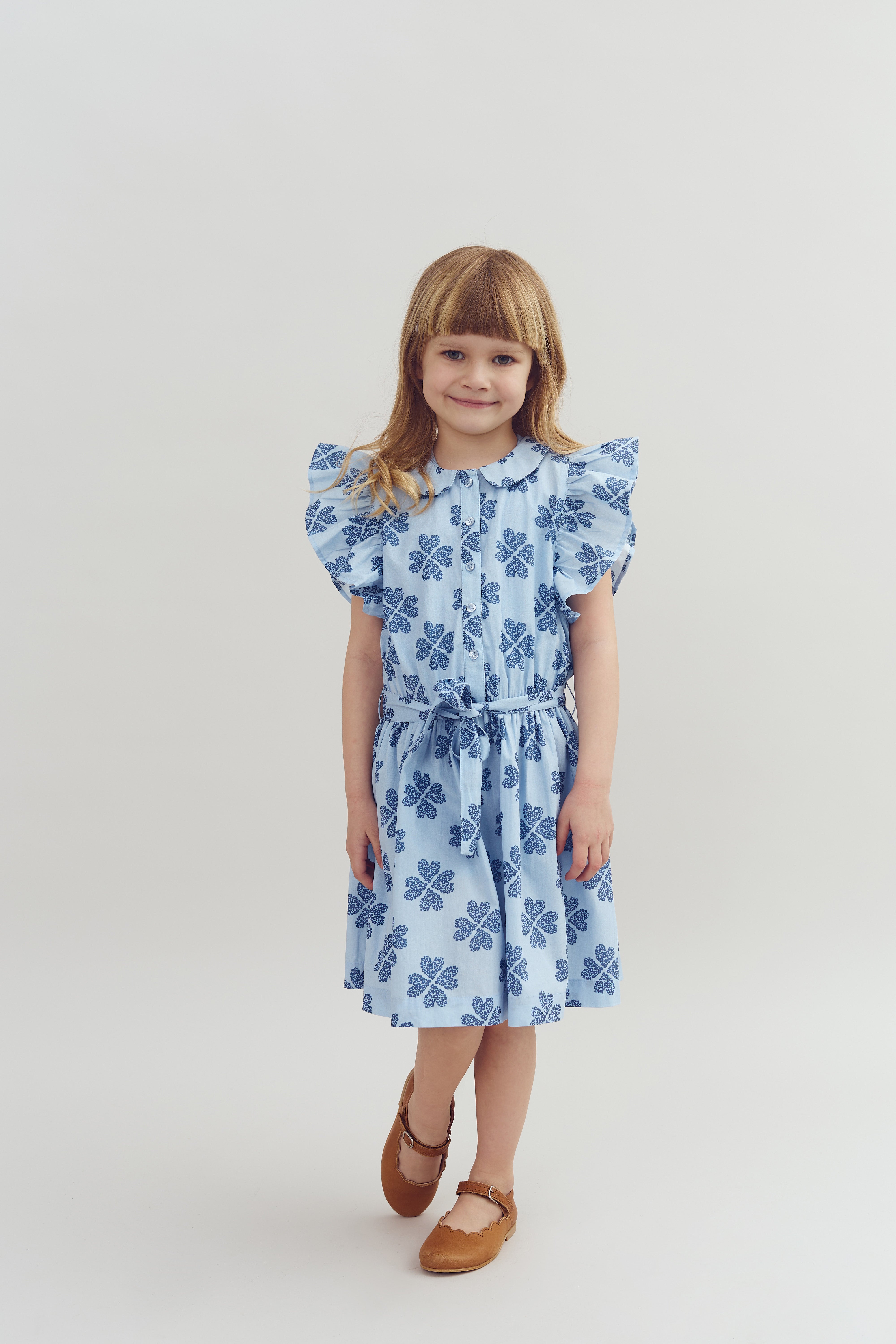A young child stands smiling, wearing a light blue dress with blue floral patterns and brown shoes, against a plain white background.