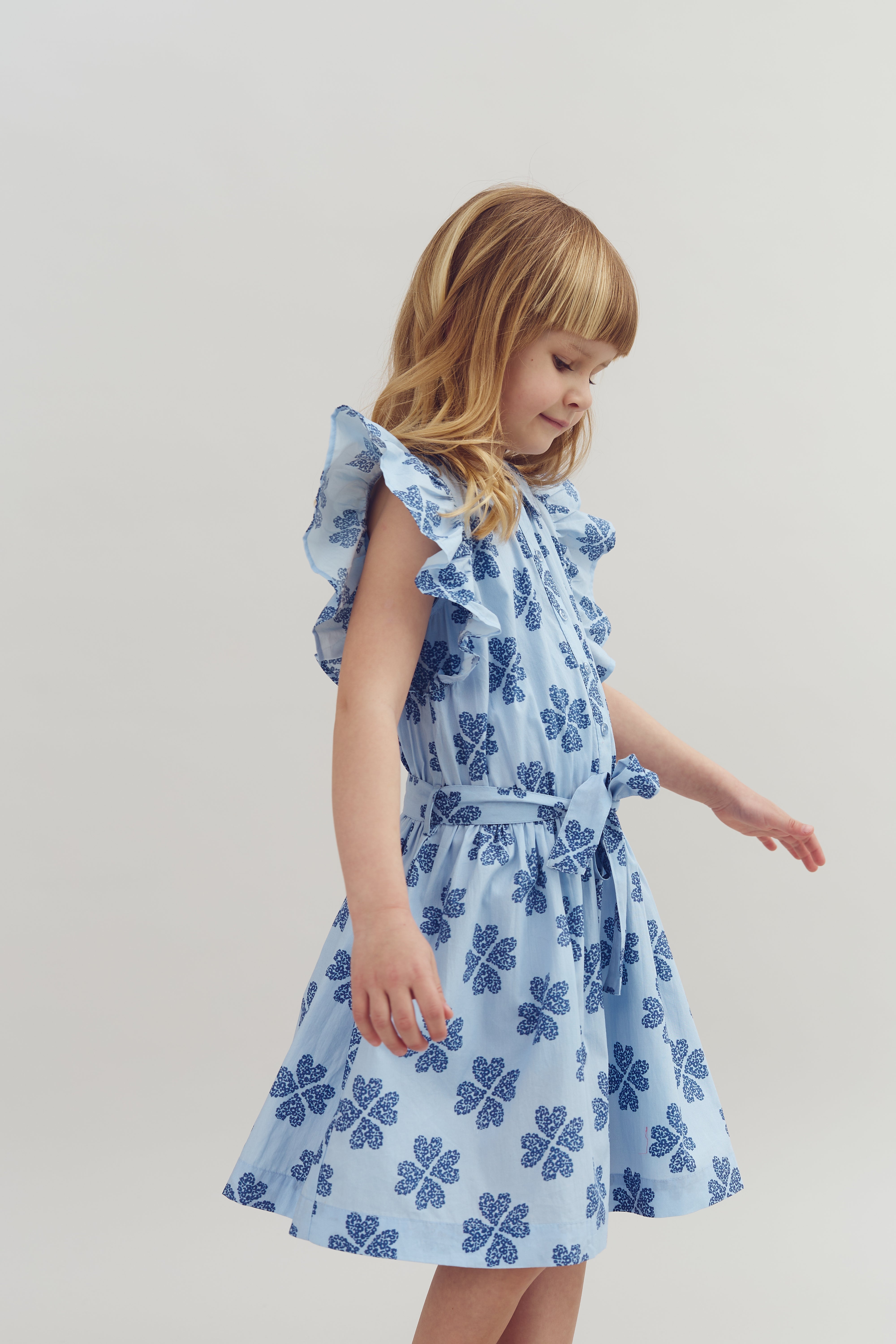 A young girl in a blue dress with dark blue patterns is looking downward. The dress features ruffled sleeves and a matching belt, set against a plain white background.