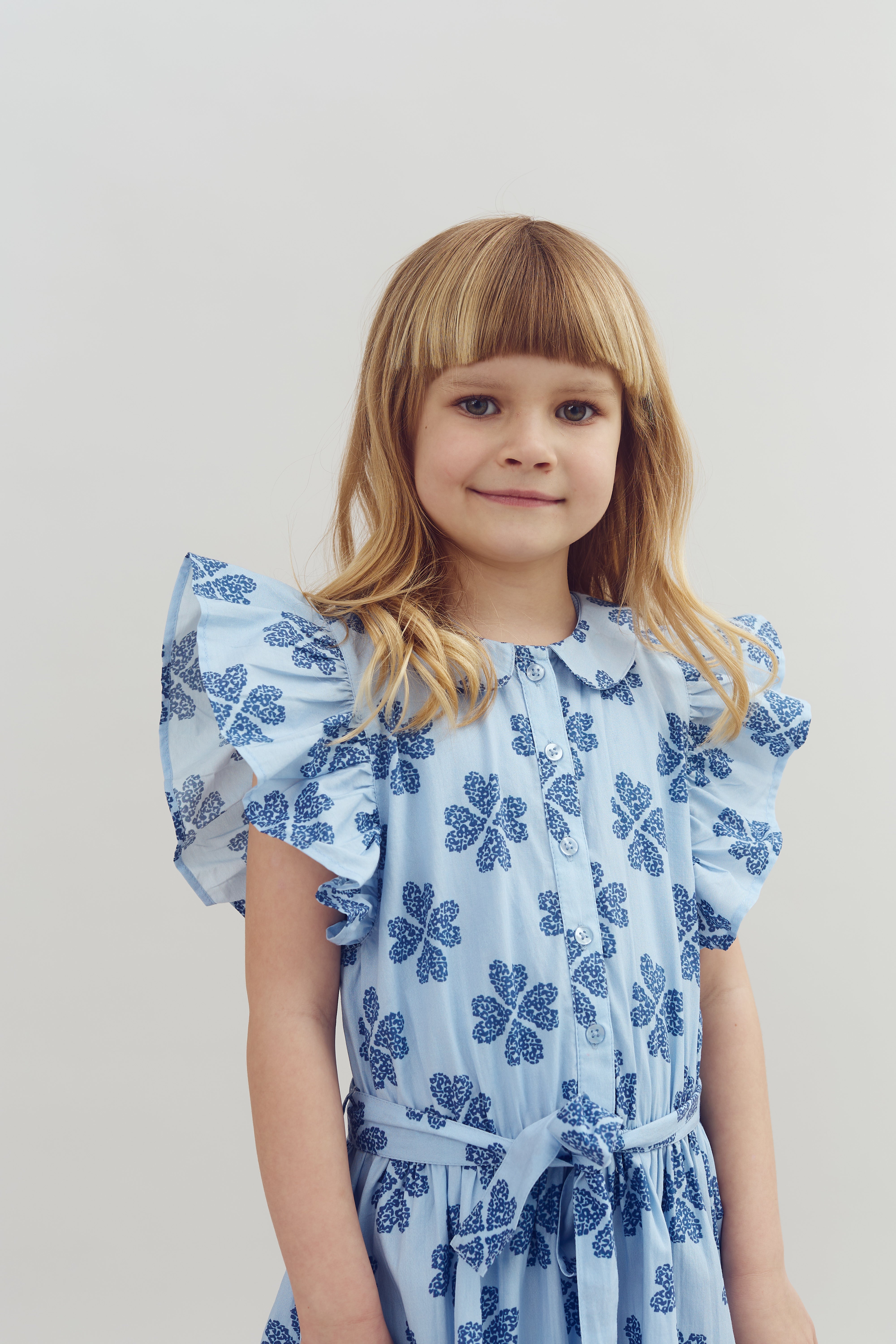 A child stands with a gentle smile, wearing a light blue dress adorned with dark blue floral patterns, against a plain, light-colored background.