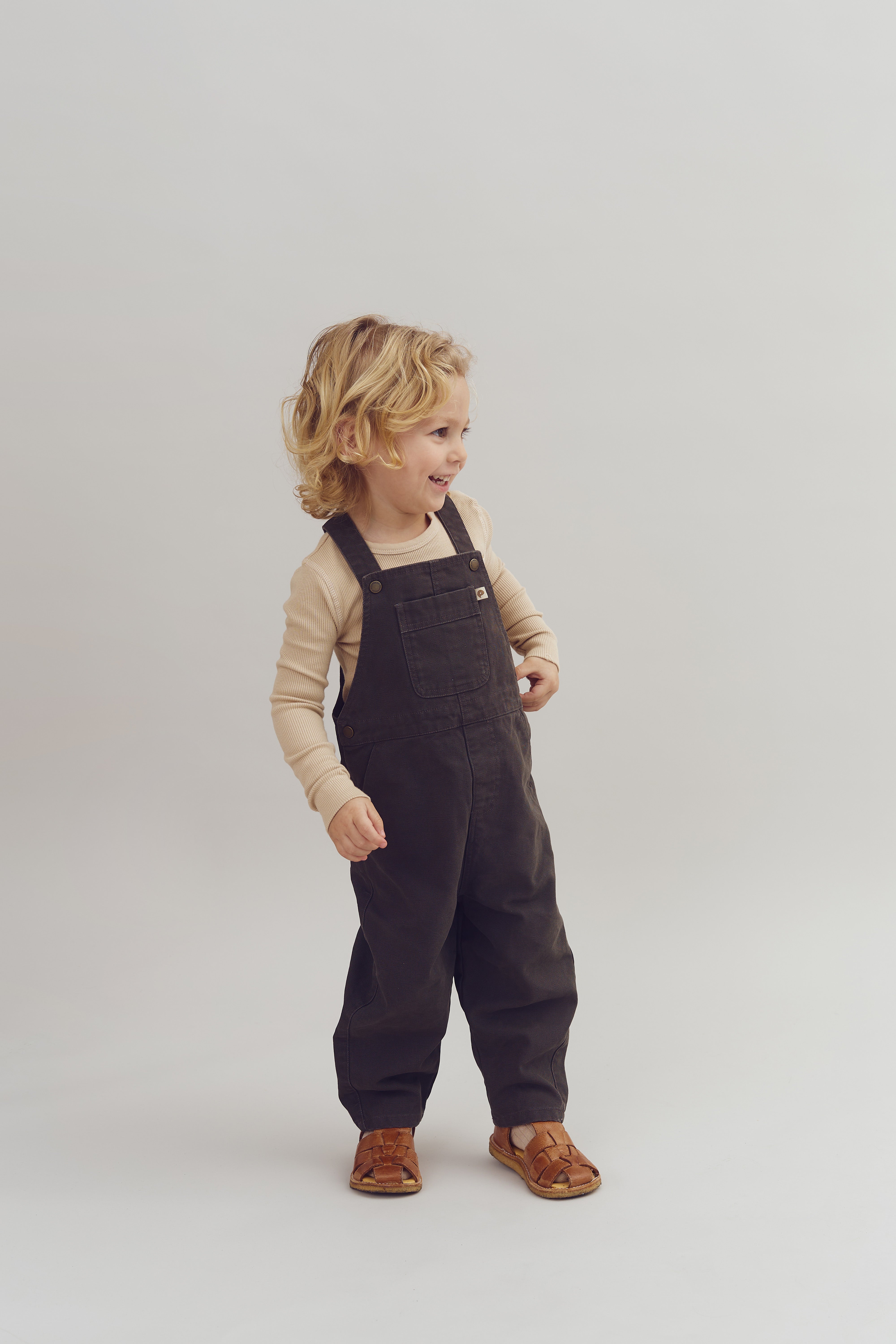 A child with curly hair stands smiling, wearing a beige long-sleeve shirt and dark overalls. They are in a brightly lit, neutral-colored studio.
