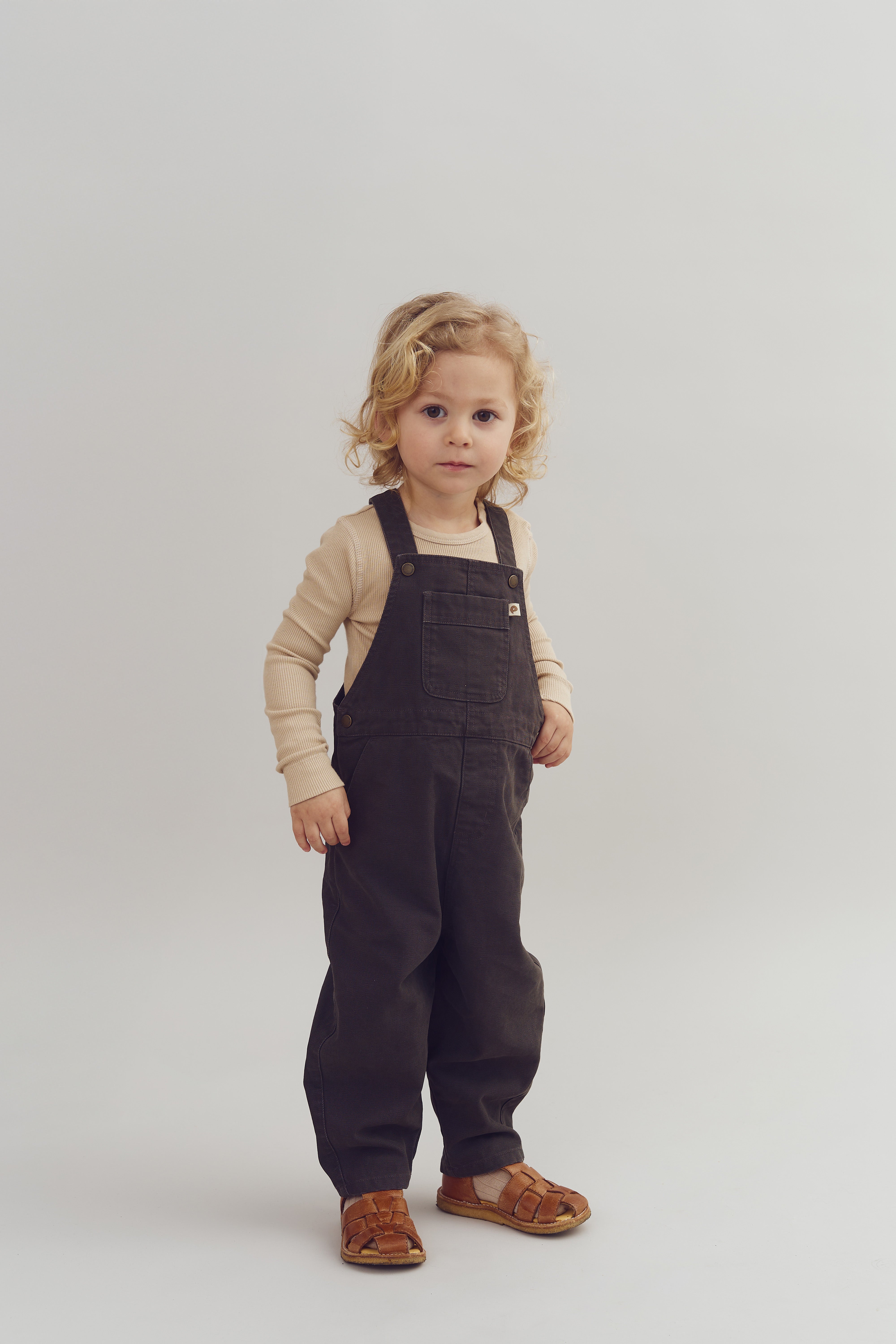 A young child stands, wearing dark overalls and a beige shirt, on a plain light gray background, looking directly at the camera.