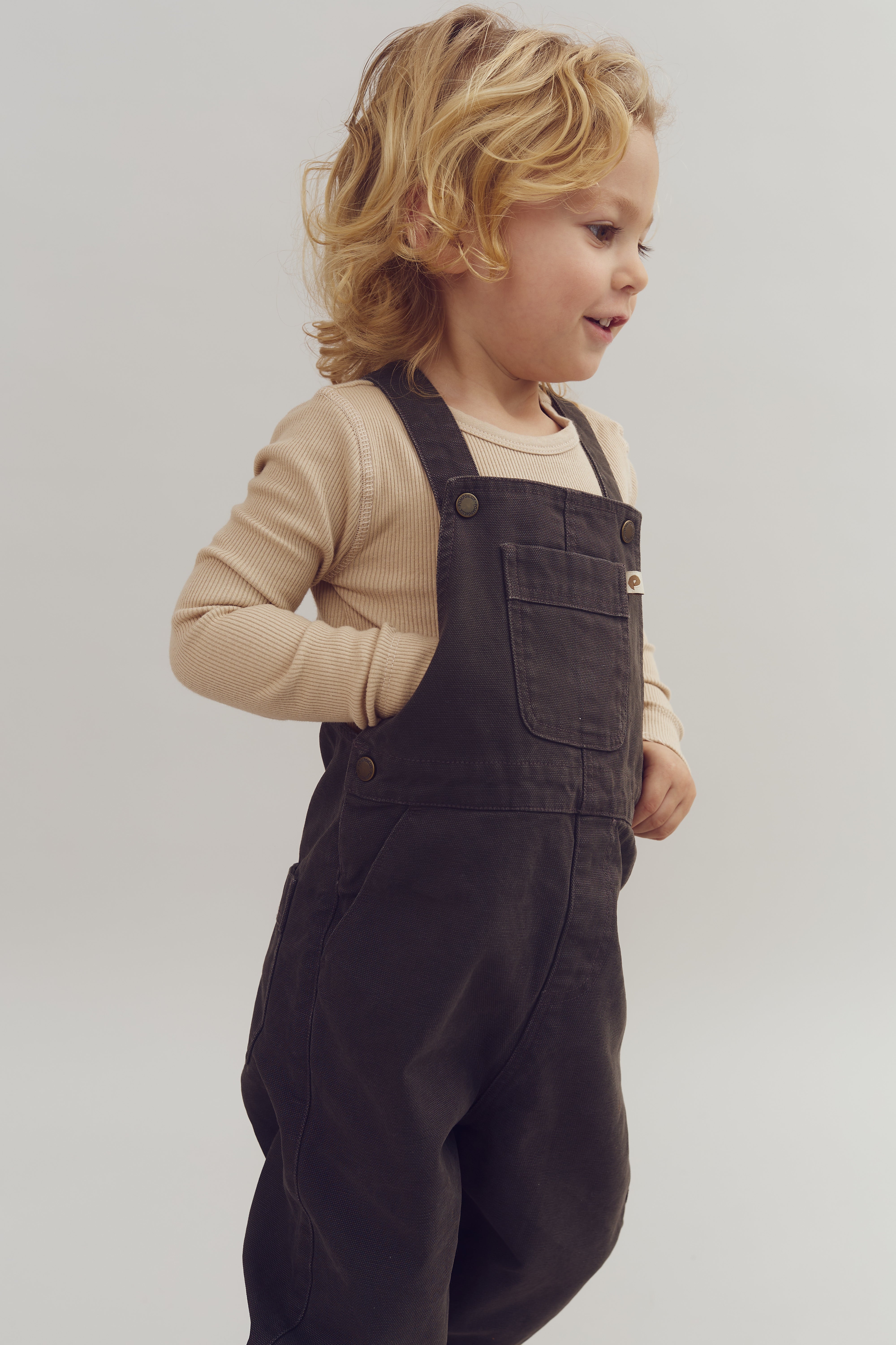 Child wearing dark overalls and a beige long-sleeve shirt is standing against a plain background, hands in pockets, smiling slightly and gazing to the side.