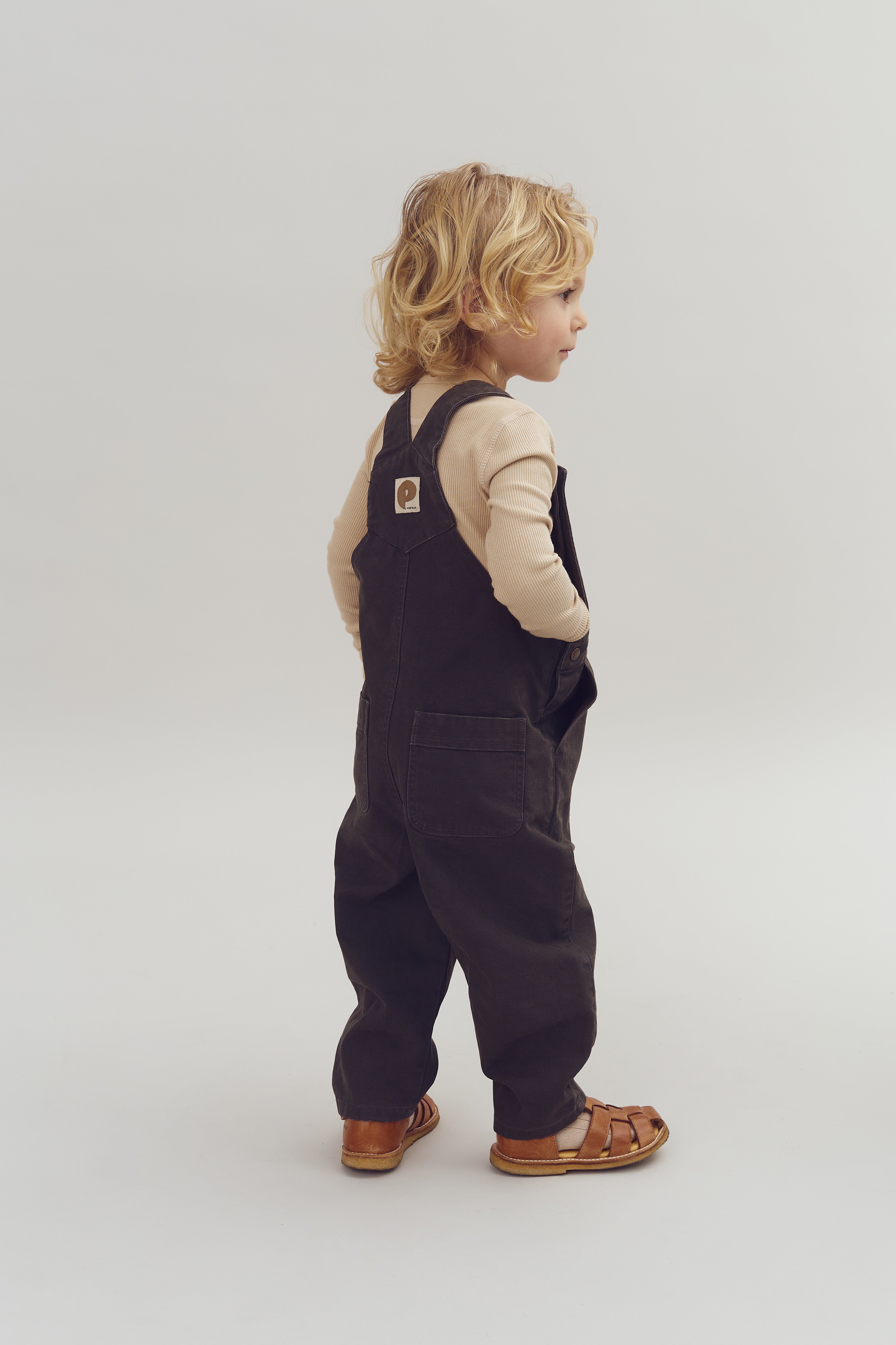 A child with curly hair stands in profile, wearing a beige shirt, dark overalls, and brown sandals. The background is plain and light-colored.