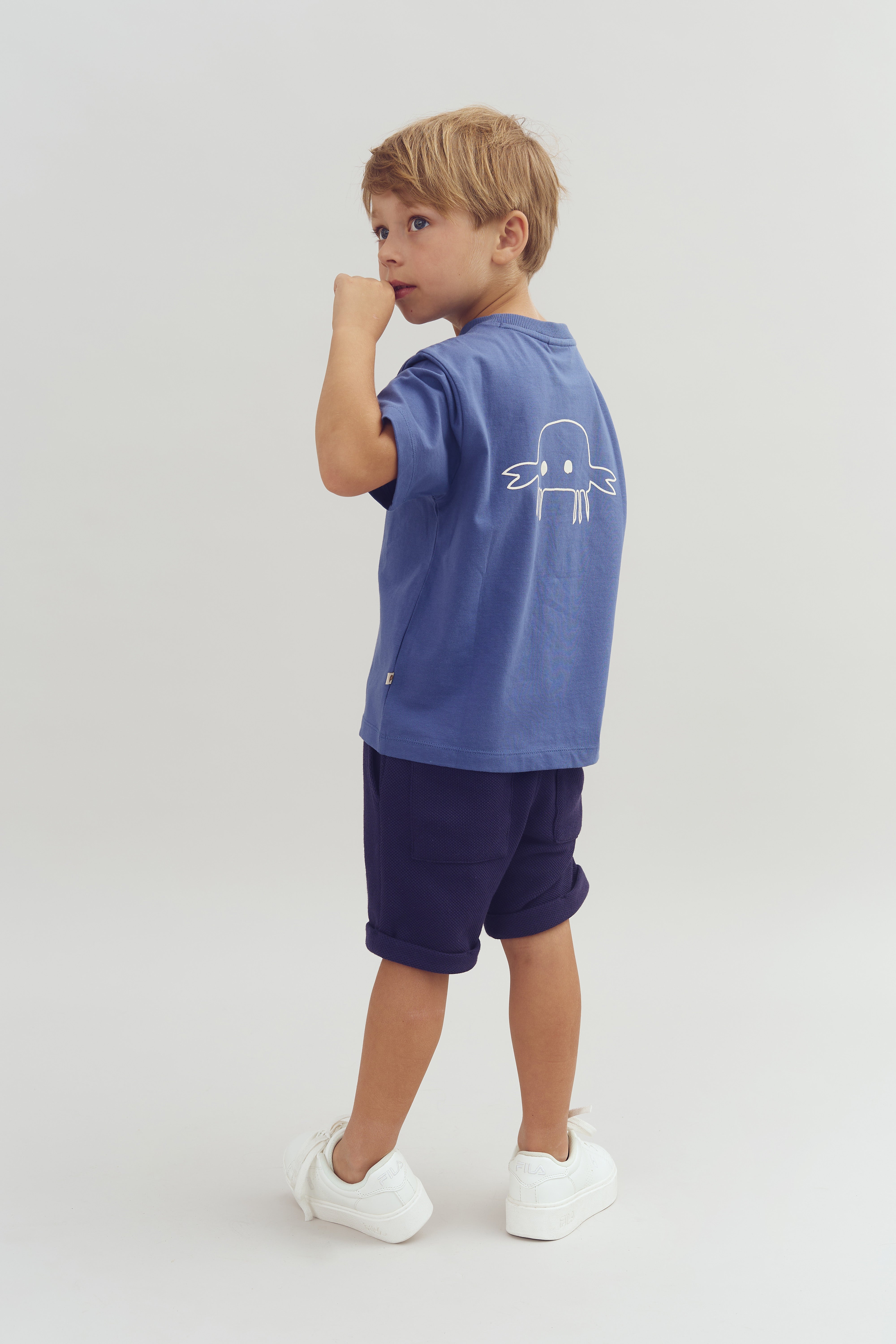 A child wearing a blue t-shirt with a white illustrated creature, navy shorts, and white sneakers stands on a plain light background, looking over their shoulder while lightly touching their mouth.