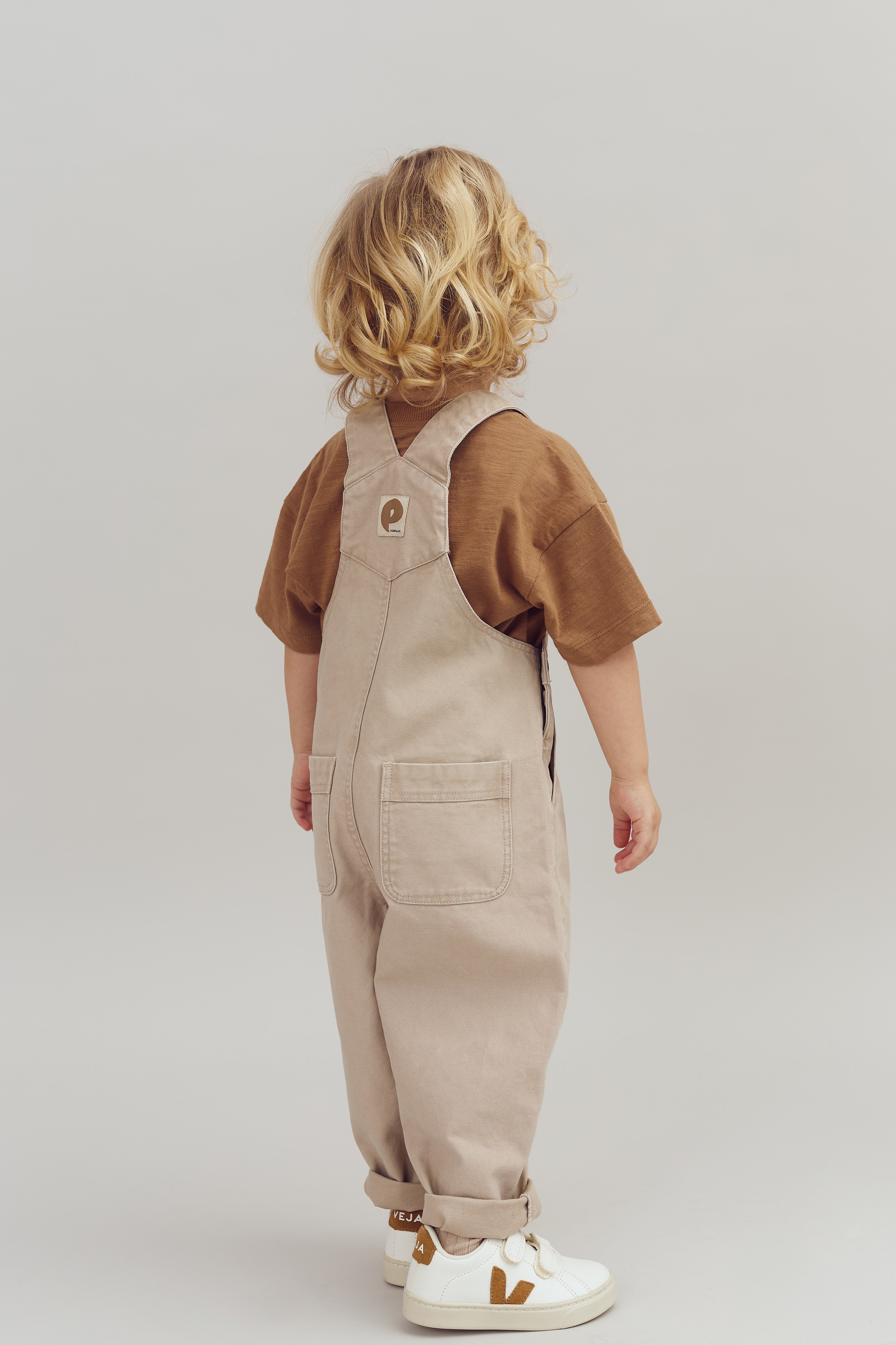 A young child with curly hair stands wearing beige overalls and brown shirt, facing away. The setting is a simple, neutral background, and their sneakers read "VEJA" on the back.