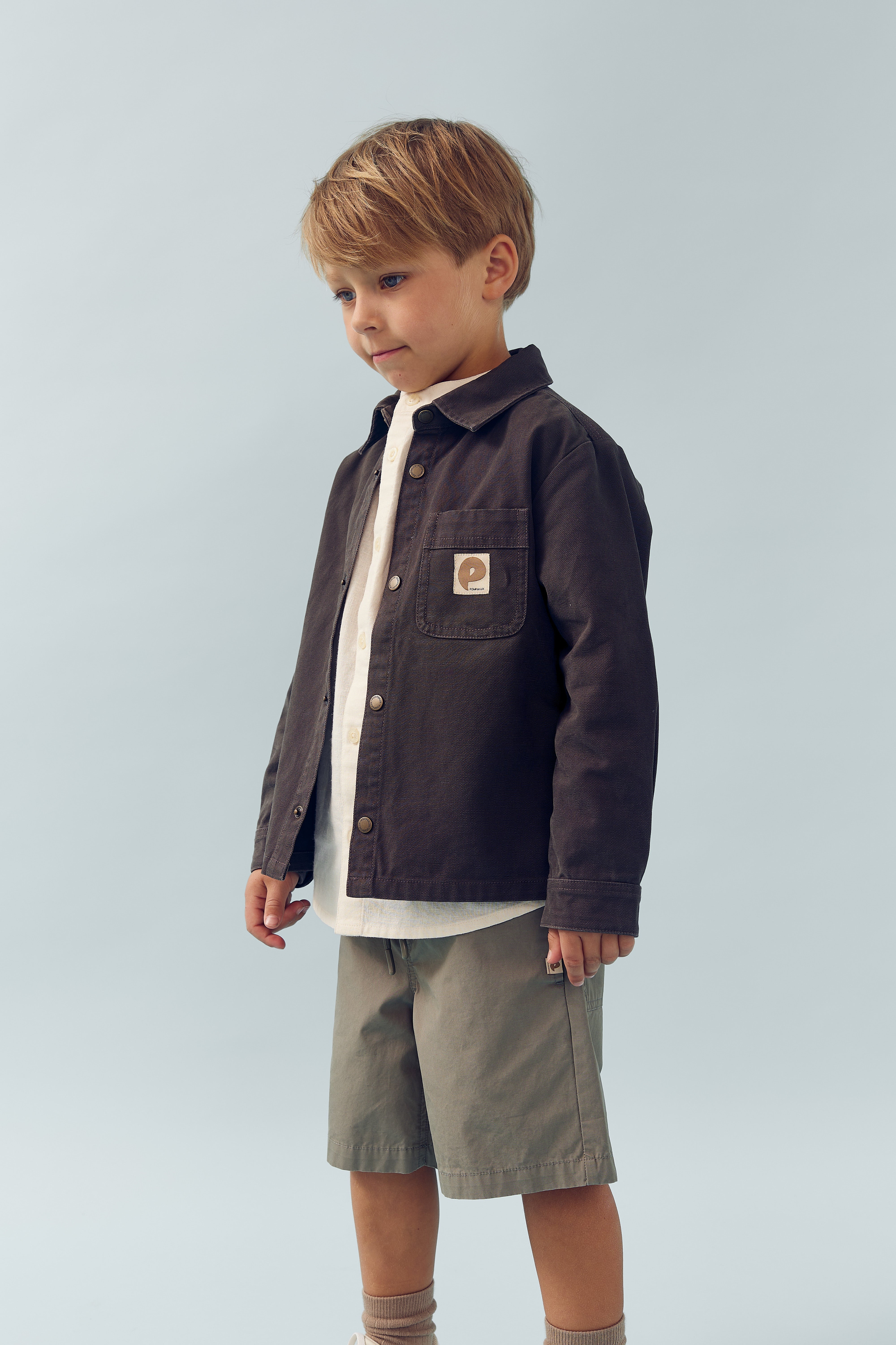 A young child stands, wearing a dark jacket with a patch and shorts, in front of a plain light blue background.