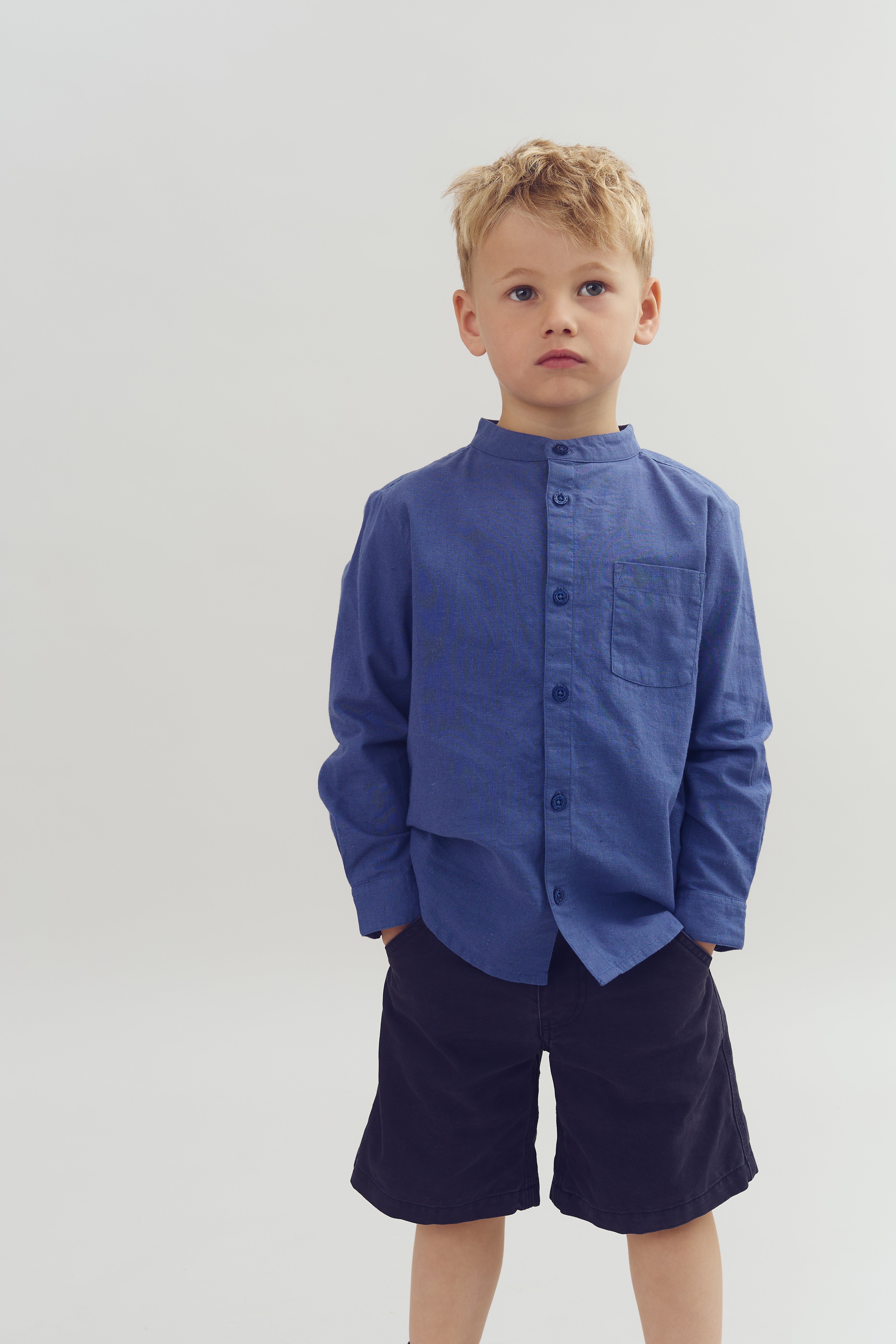 A young child stands with hands in pockets, wearing a blue button-up shirt and dark shorts, set against a neutral background.