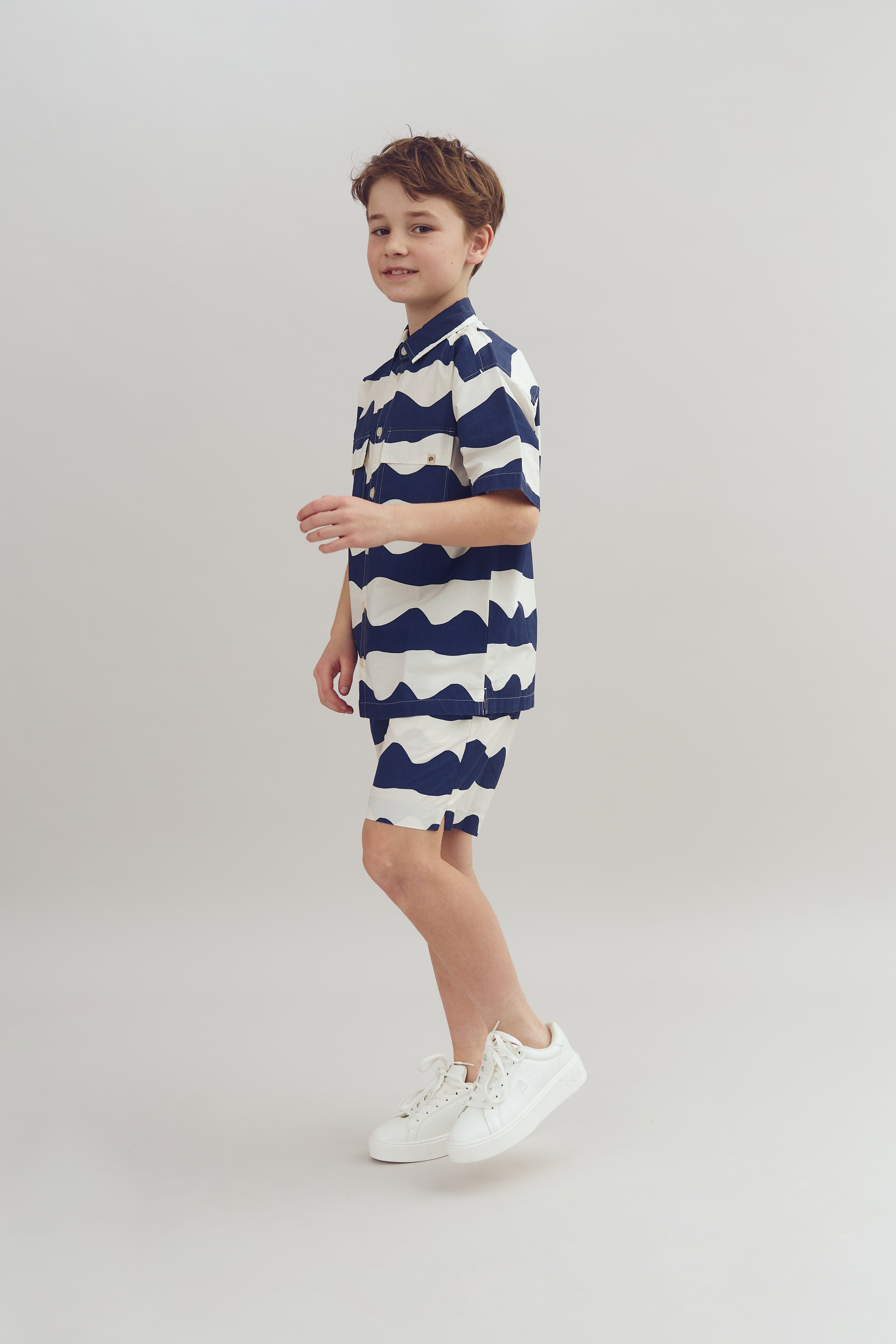 A smiling child wearing a blue-and-white wave-patterned shirt and shorts stands casually in a minimalist, light-colored studio setting.