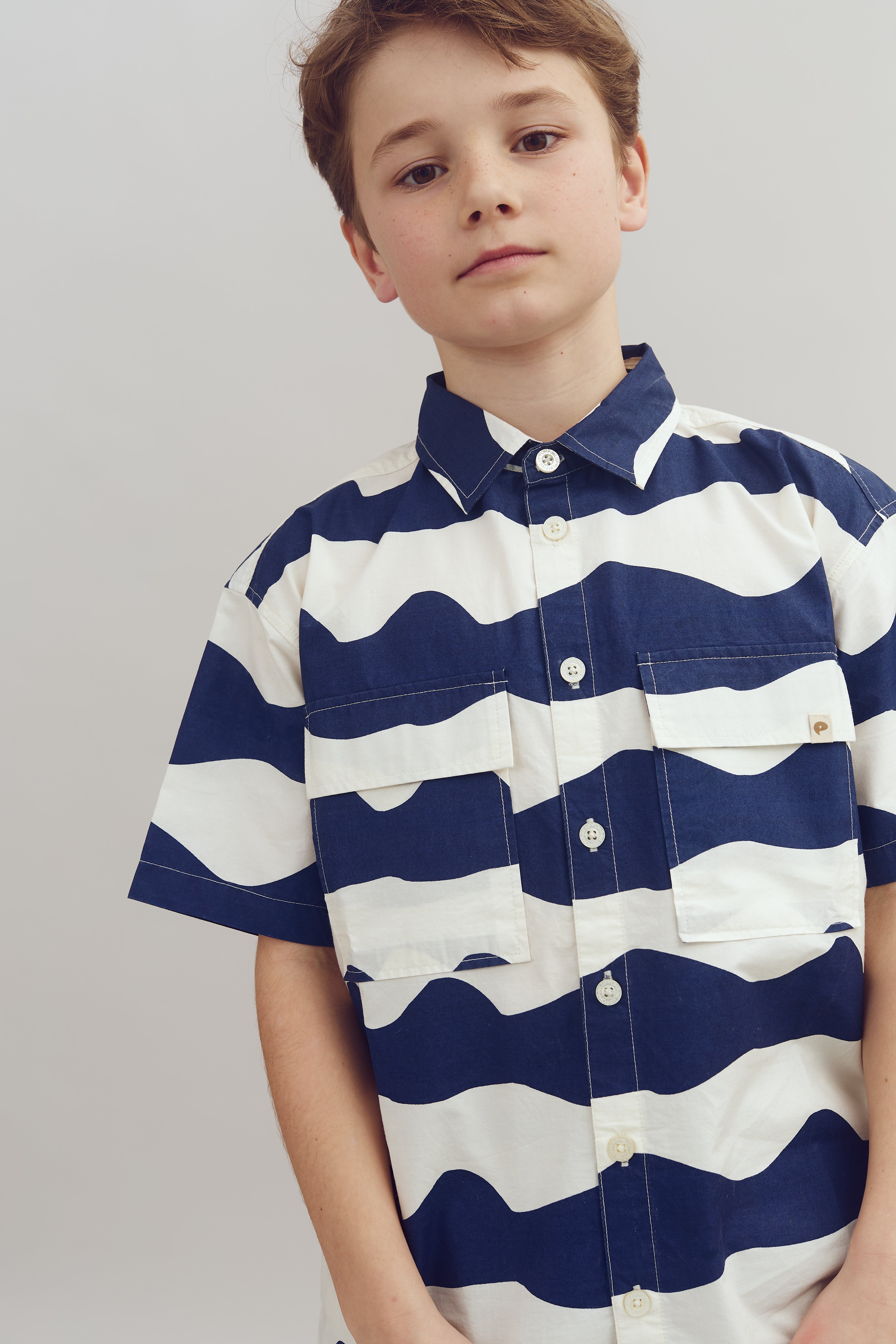 A person wearing a short-sleeved shirt with bold blue and white wave-like stripes stands against a plain gray background, looking directly at the camera.