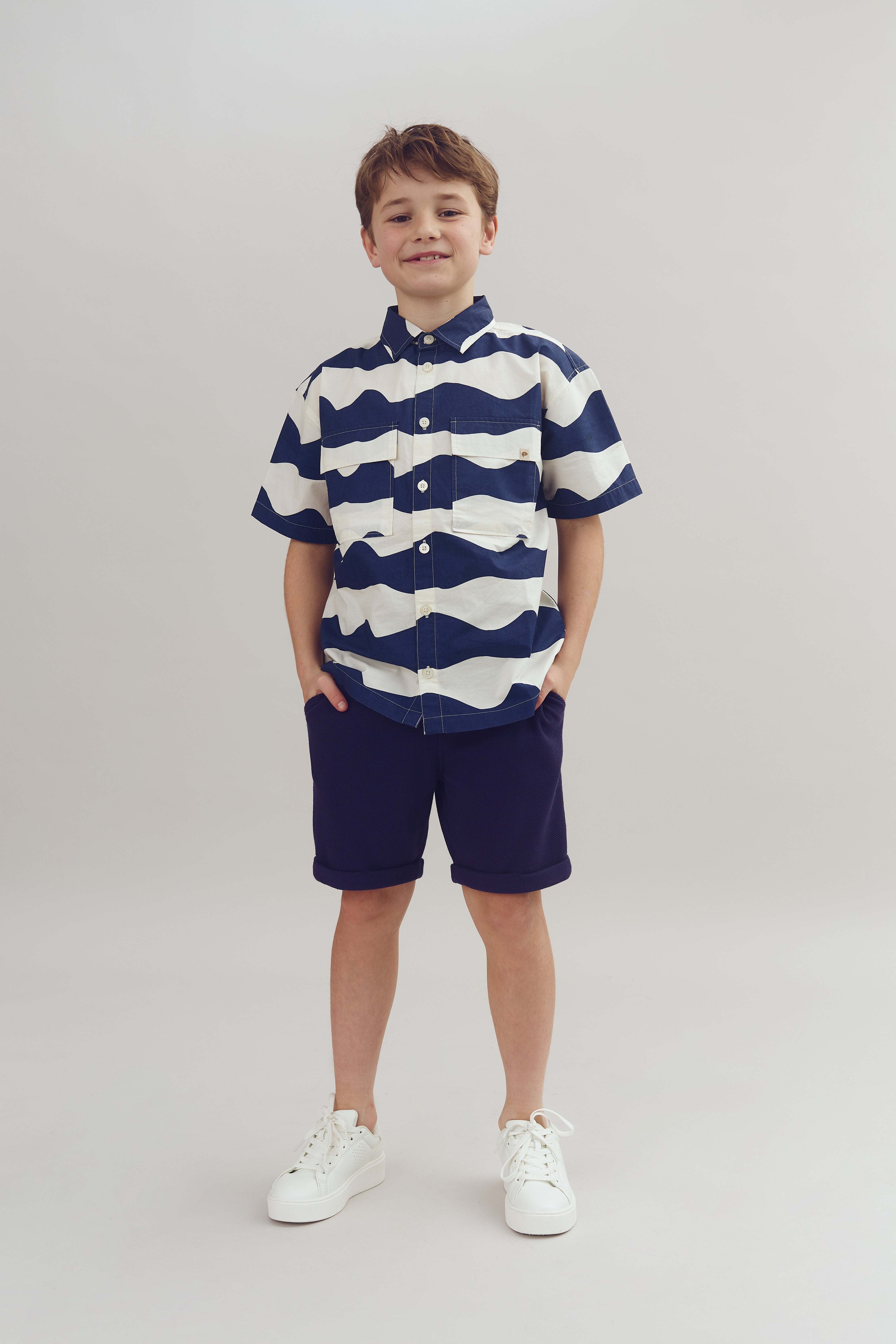 A child stands smiling, hands in pockets, wearing a wavy-patterned shirt, shorts, and white sneakers, against a plain beige background.