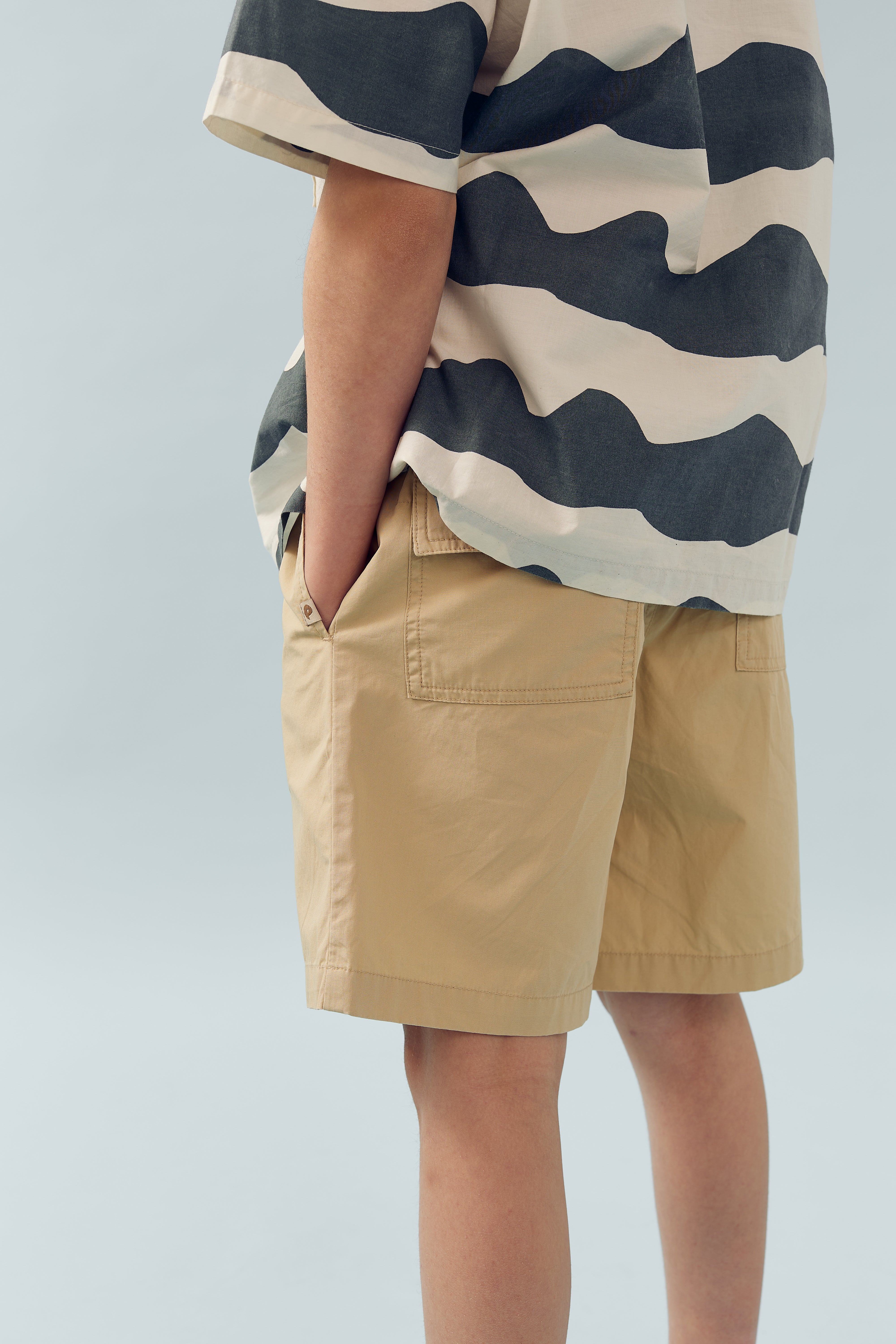 A person stands with hands in pockets, wearing a striped shirt and beige shorts. The background is a plain light blue, providing a neutral setting.