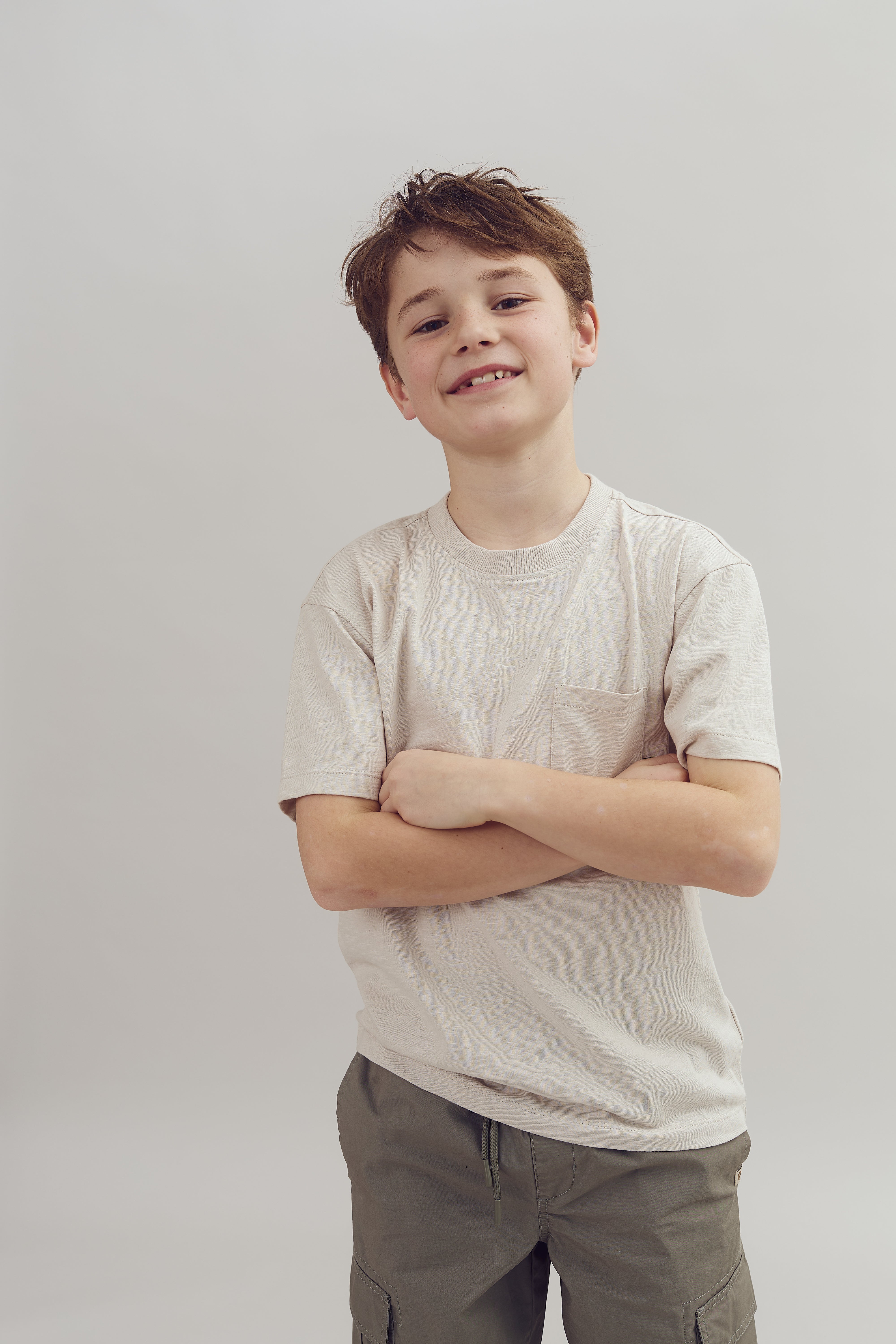A young boy stands confidently with arms crossed, wearing a beige t-shirt and gray pants, against a plain gray background.
