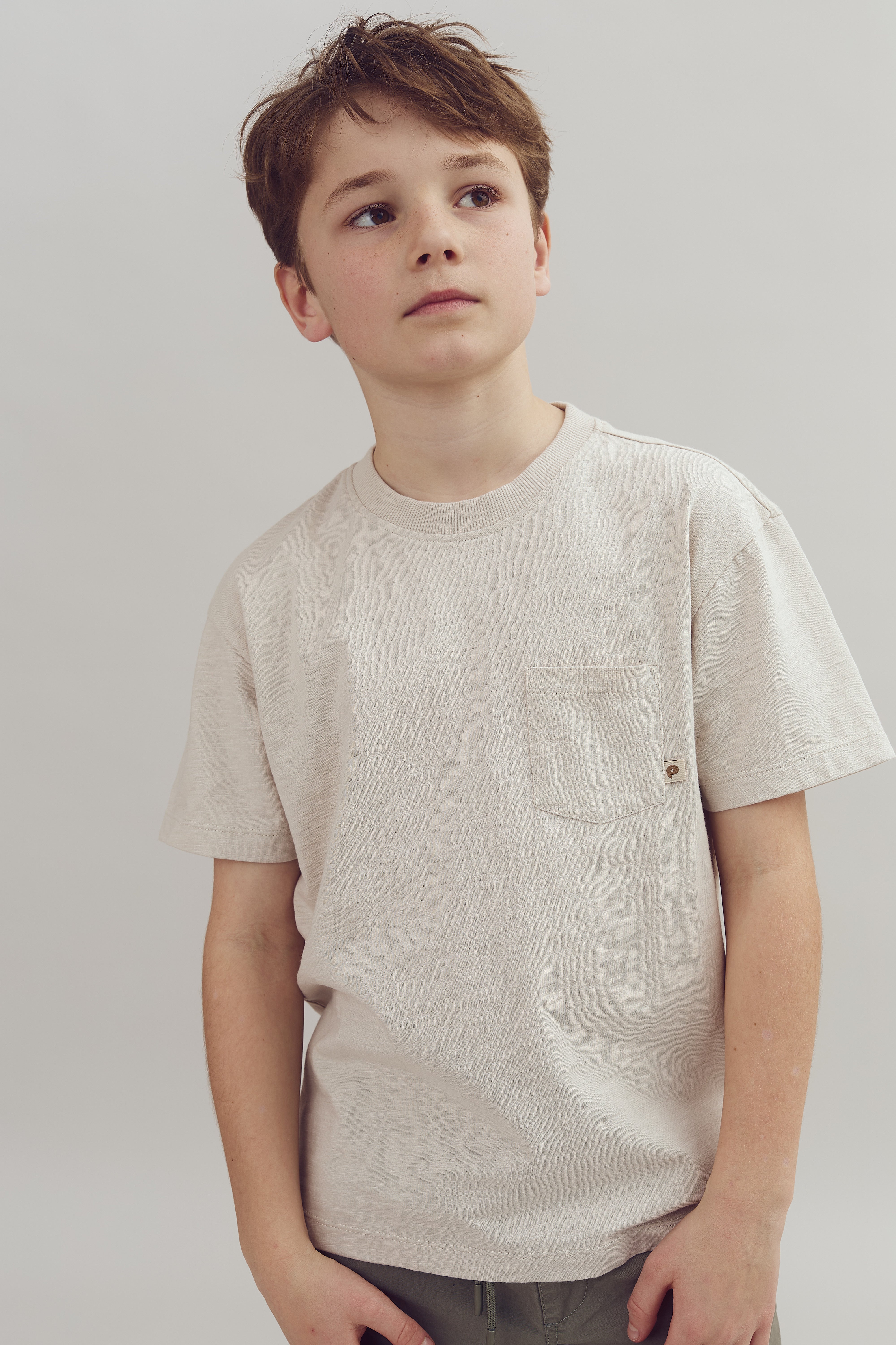 A young boy wearing a textured, light-colored t-shirt with a chest pocket stands looking upwards thoughtfully against a plain, light background.