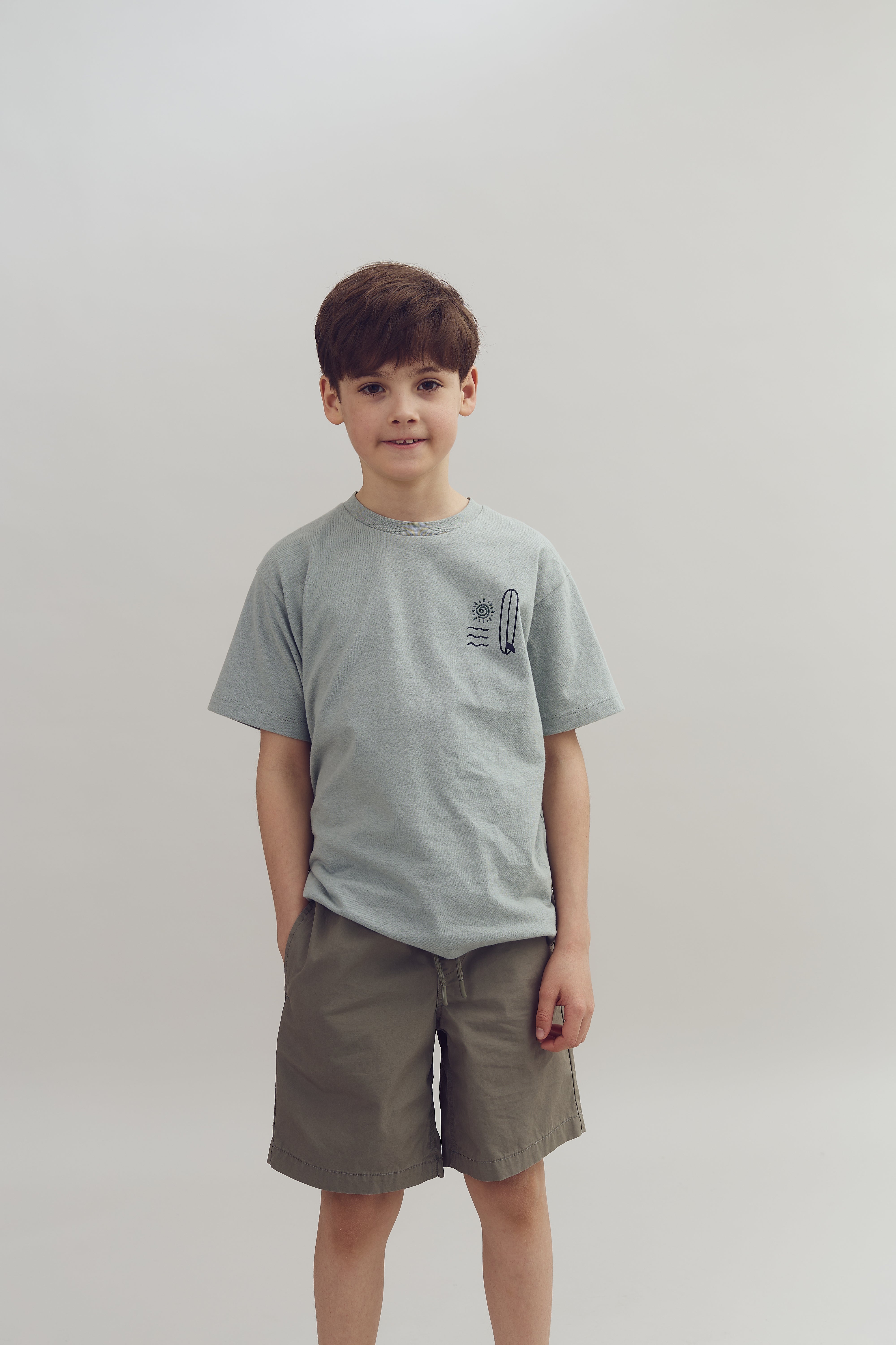 A young boy stands facing the viewer, wearing a light gray t-shirt and brown shorts, hands in pockets, against a plain, light-colored background.