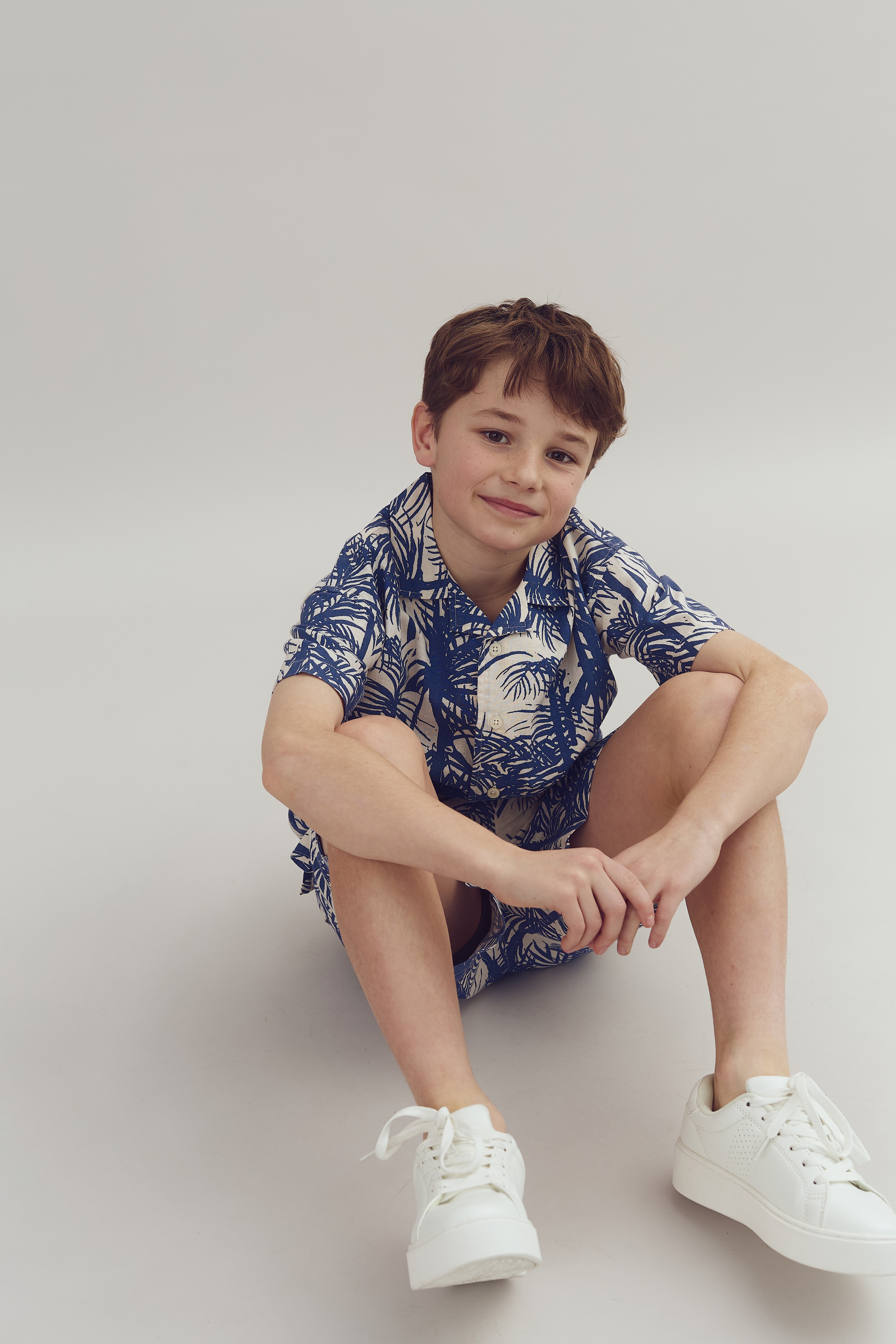 A child sits cross-legged, smiling, wearing a blue and white patterned outfit with white sneakers, against a plain, neutral background.