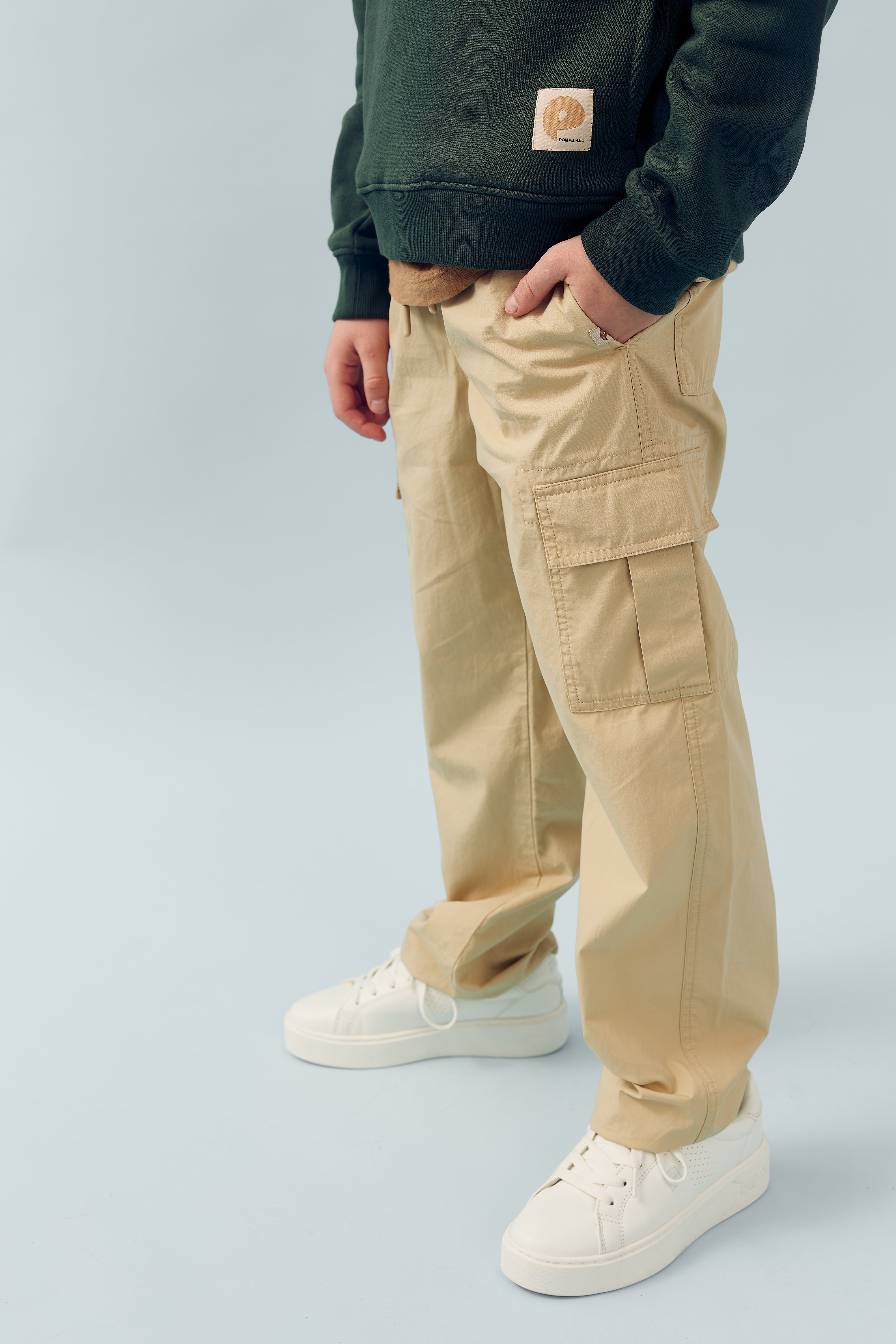 A person stands casually with one hand in the pocket of beige cargo pants, wearing a green sweater and white sneakers, against a light blue background. A patch reads "P."