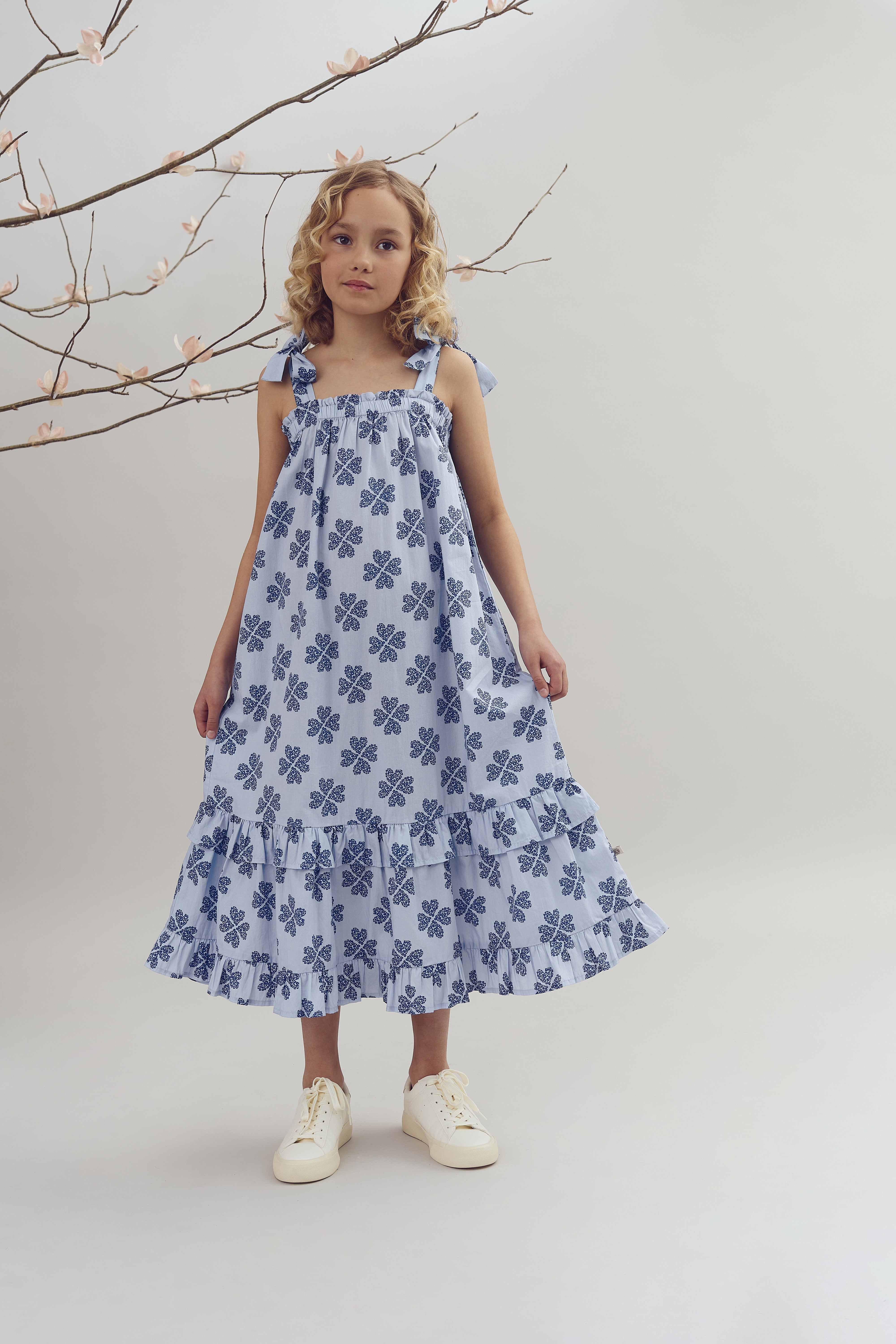 A young girl stands wearing a blue, floral-patterned dress with ruffles. She is positioned near a branch adorned with light pink blossoms against a plain background.