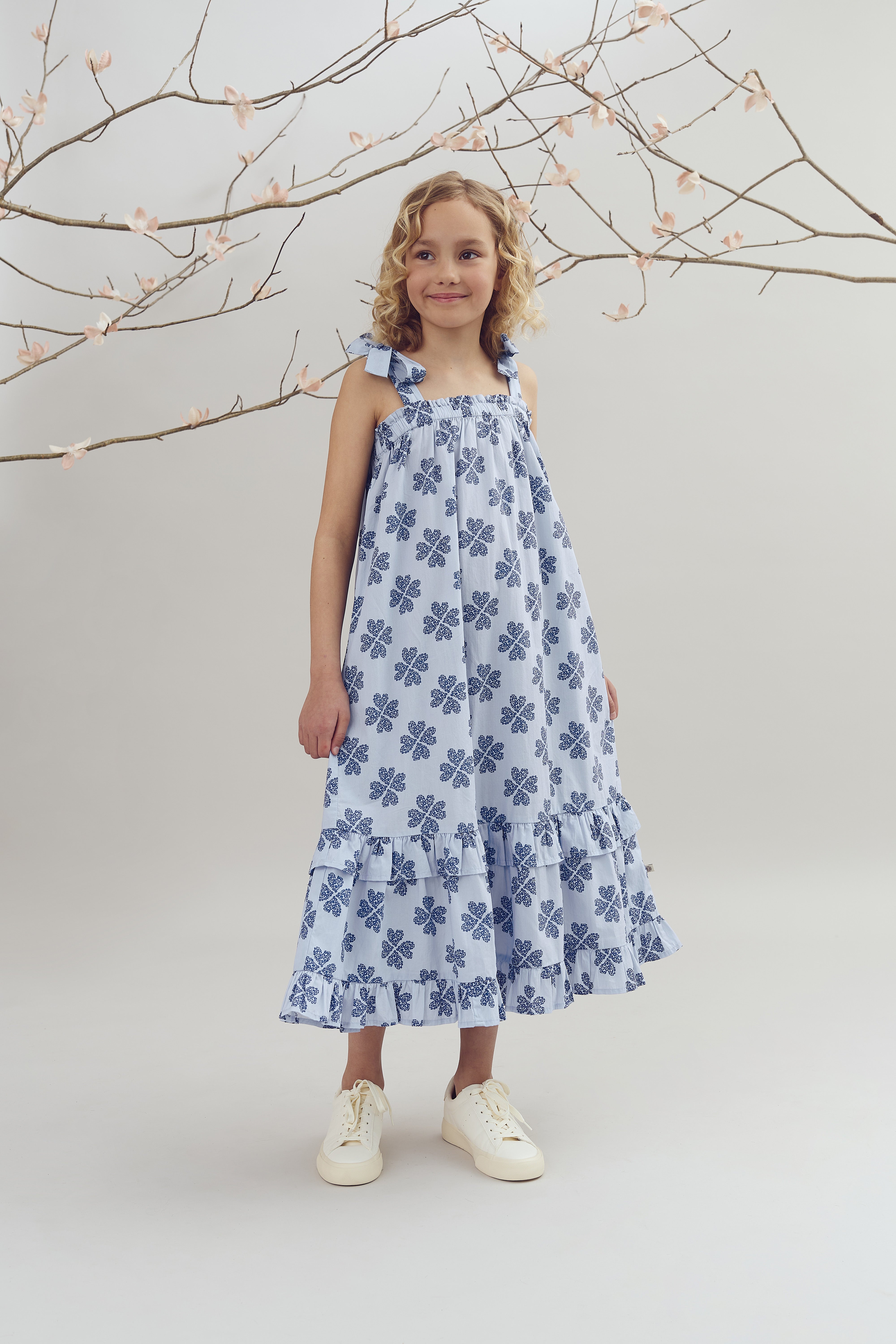 A child in a blue patterned dress stands smiling, surrounded by delicate branches with pink blossoms, set against a plain, soft-colored background.