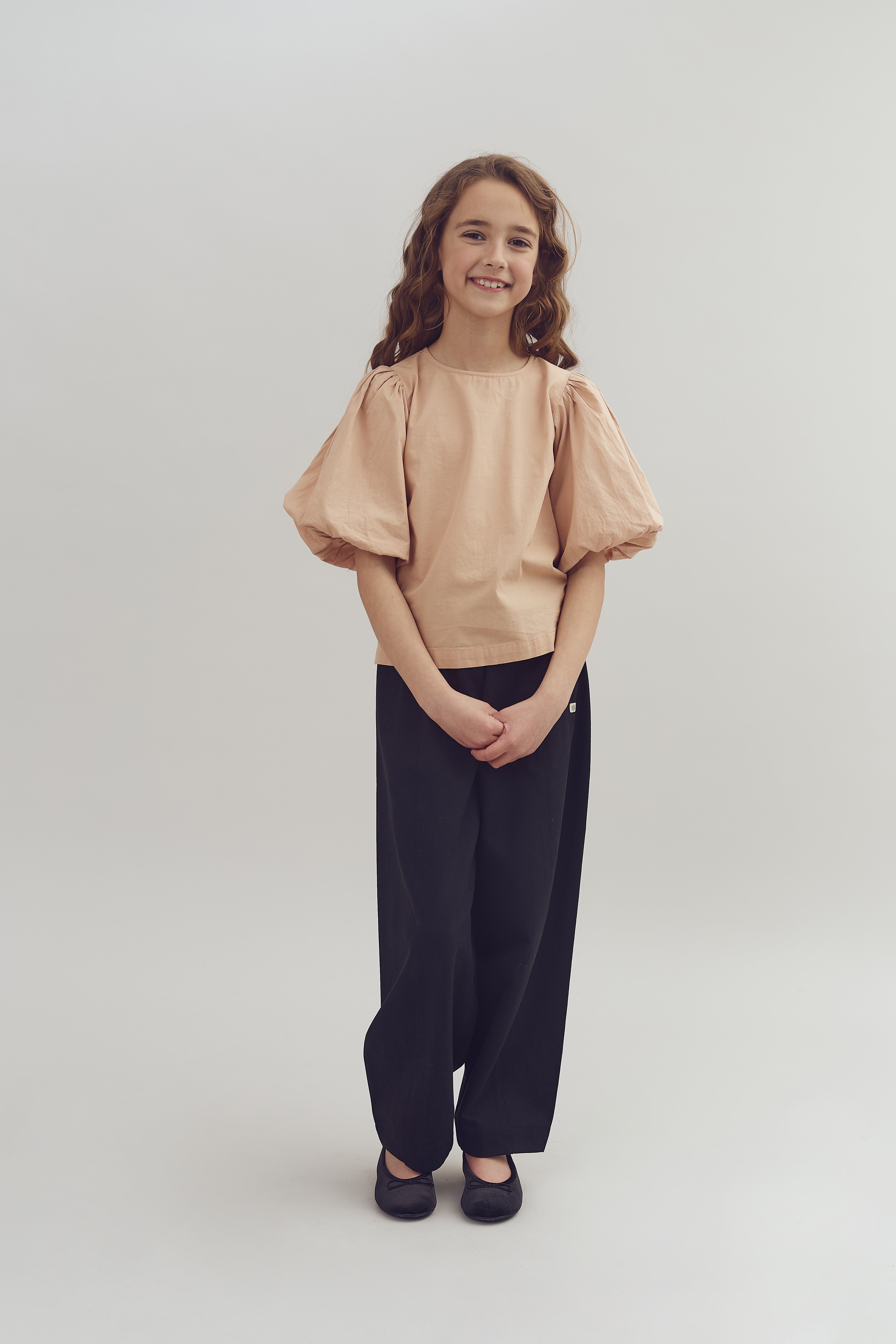 A smiling girl stands with hands clasped, wearing a peach blouse and black pants, in a neutral studio setting.