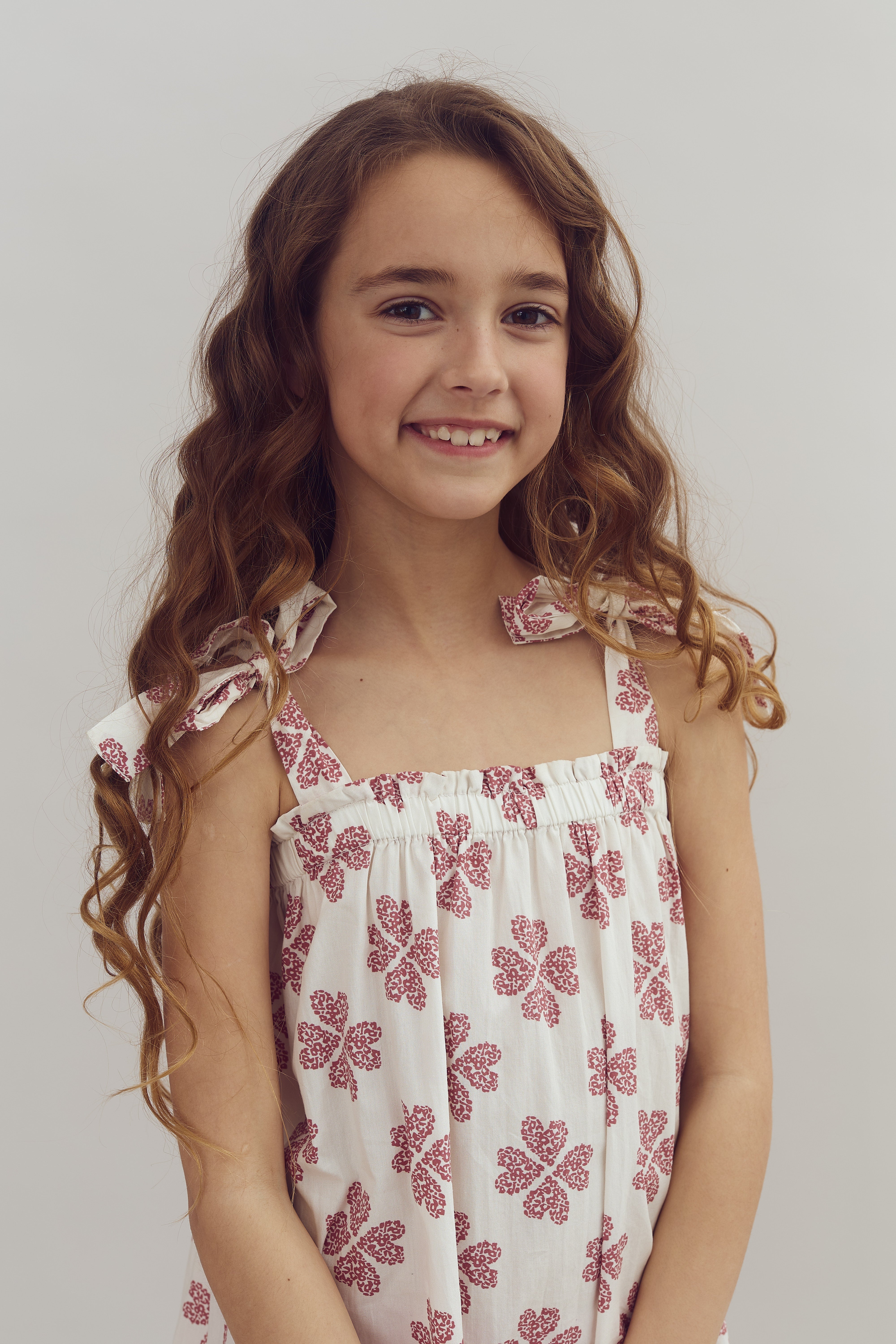 A young girl stands smiling, wearing a white dress with red floral patterns, against a plain light background.