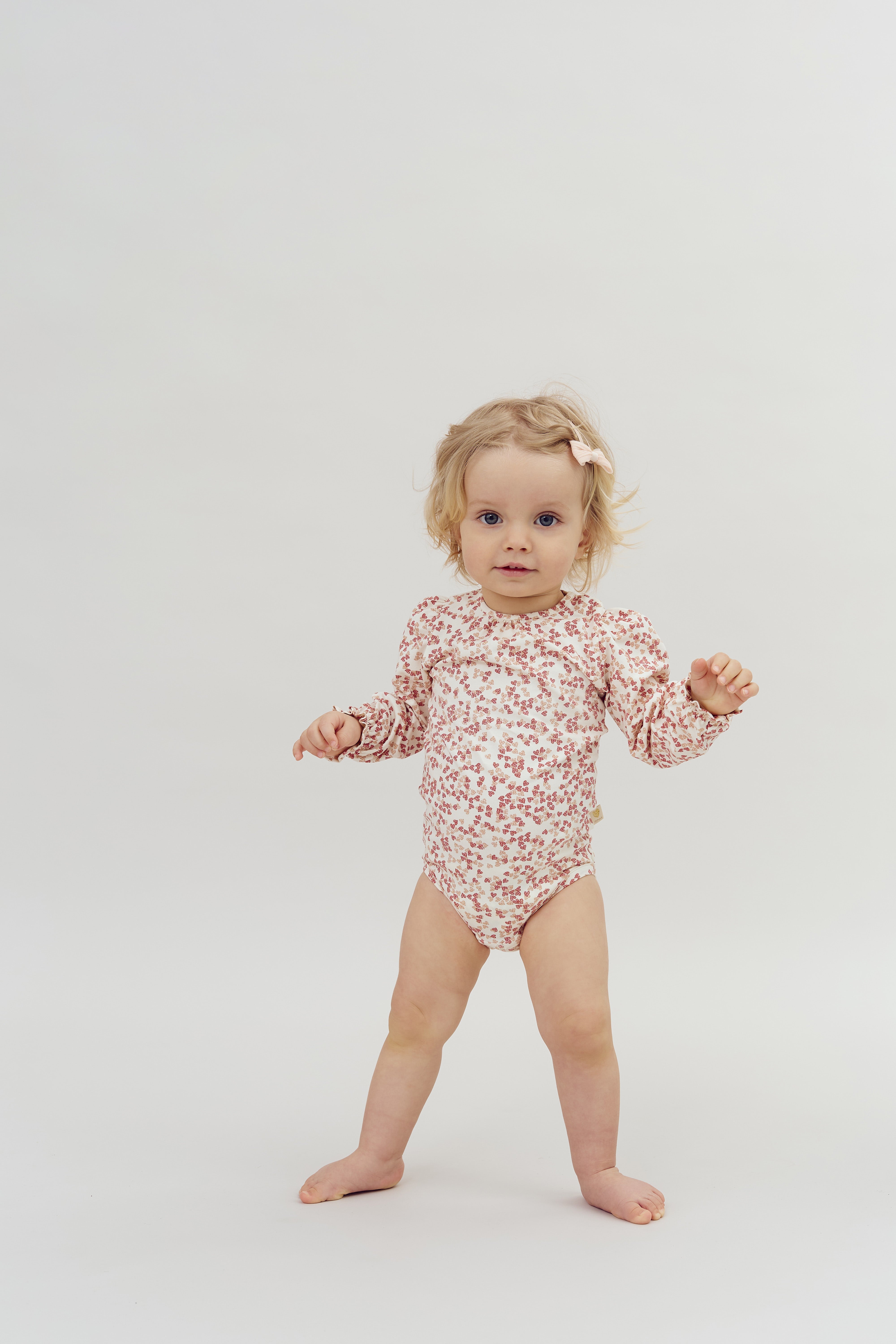 A toddler in a floral-patterned onesie stands confidently, arms outstretched, against a plain white background, with a hair bow accenting their light hair.