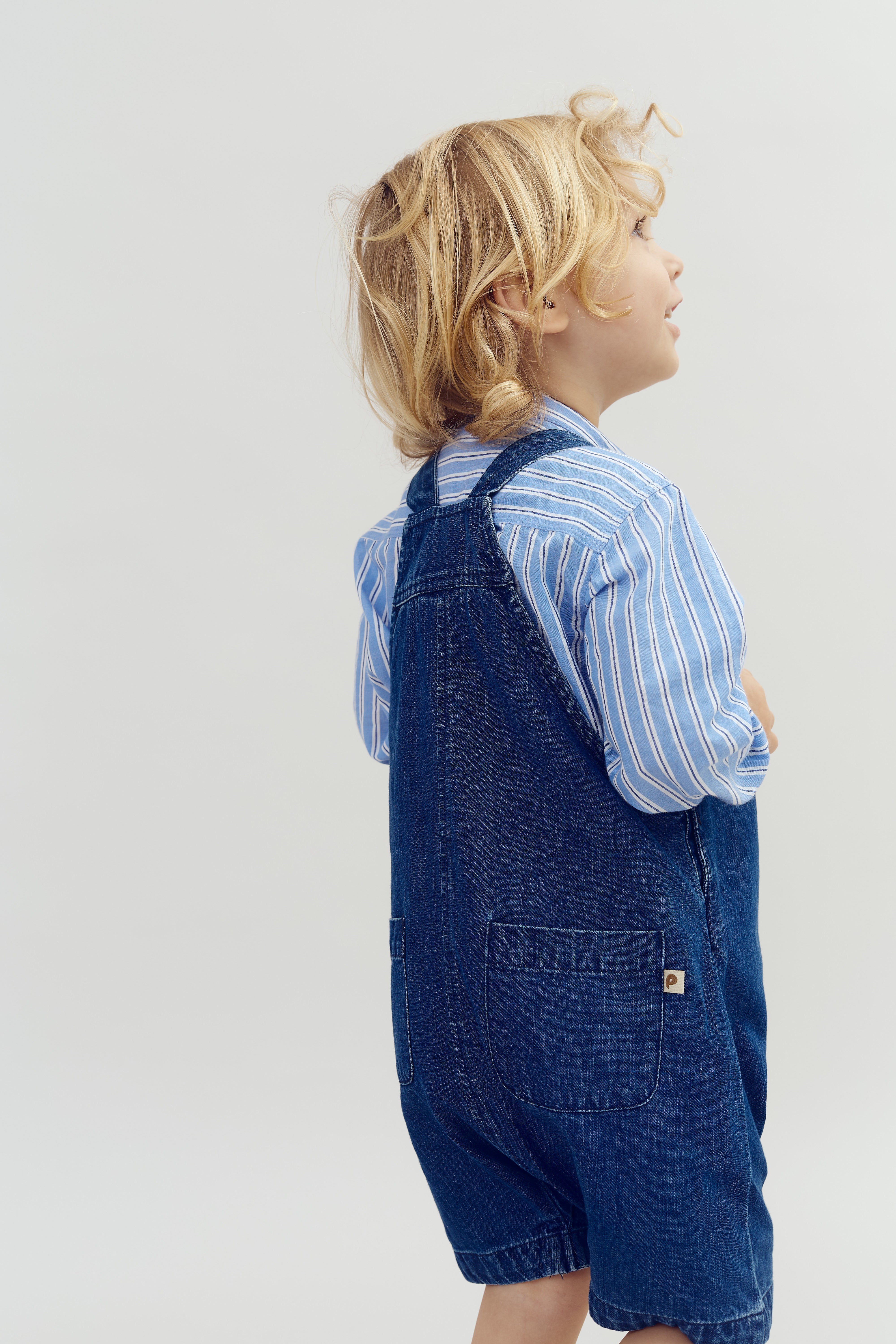 A child with blonde hair stands sideways, wearing a blue striped shirt and denim overalls, against a plain light background, looking upward with a curious expression.