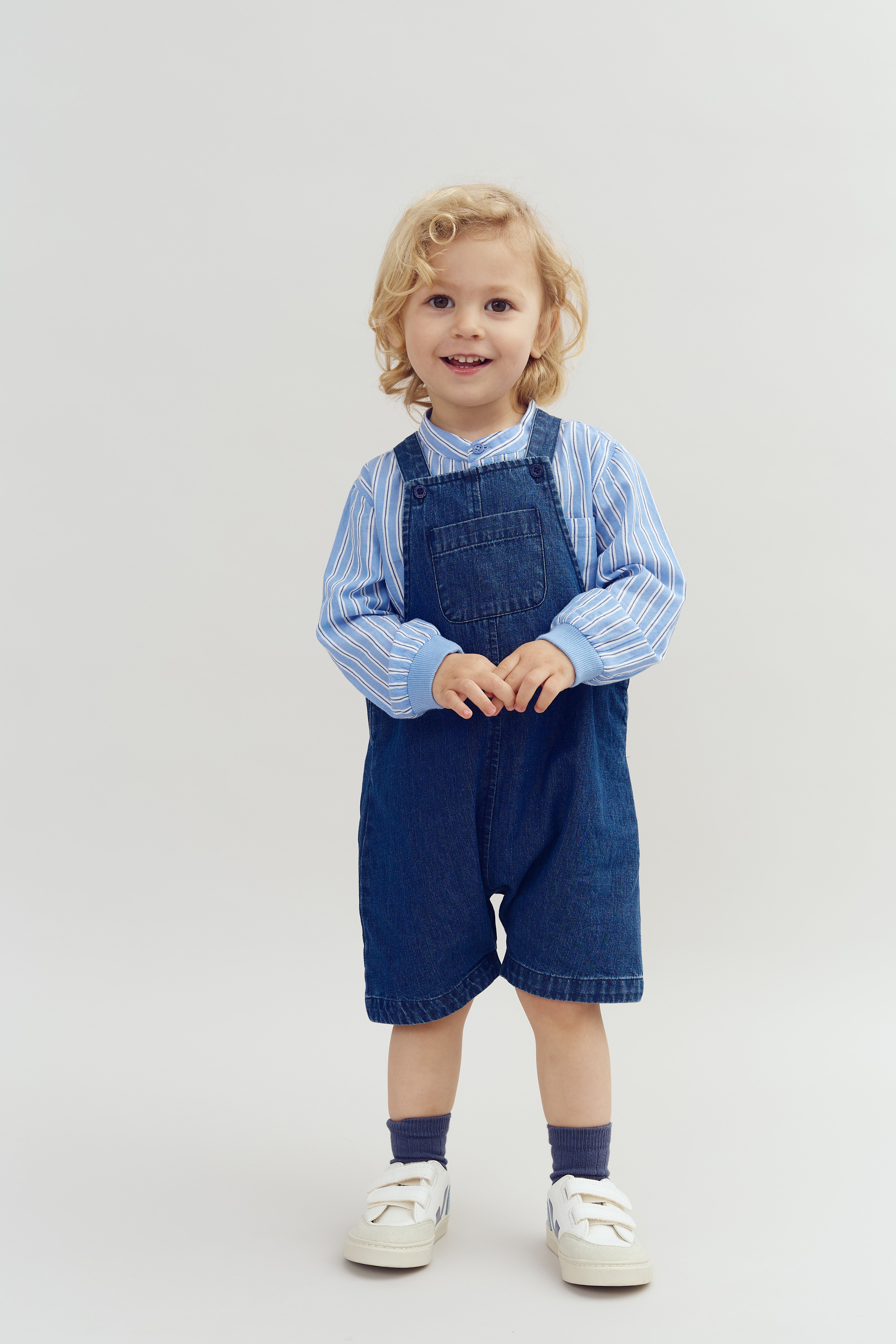 A young child stands smiling, wearing denim overalls over a blue and white striped shirt, with white sneakers, in a neutral, well-lit studio setting.