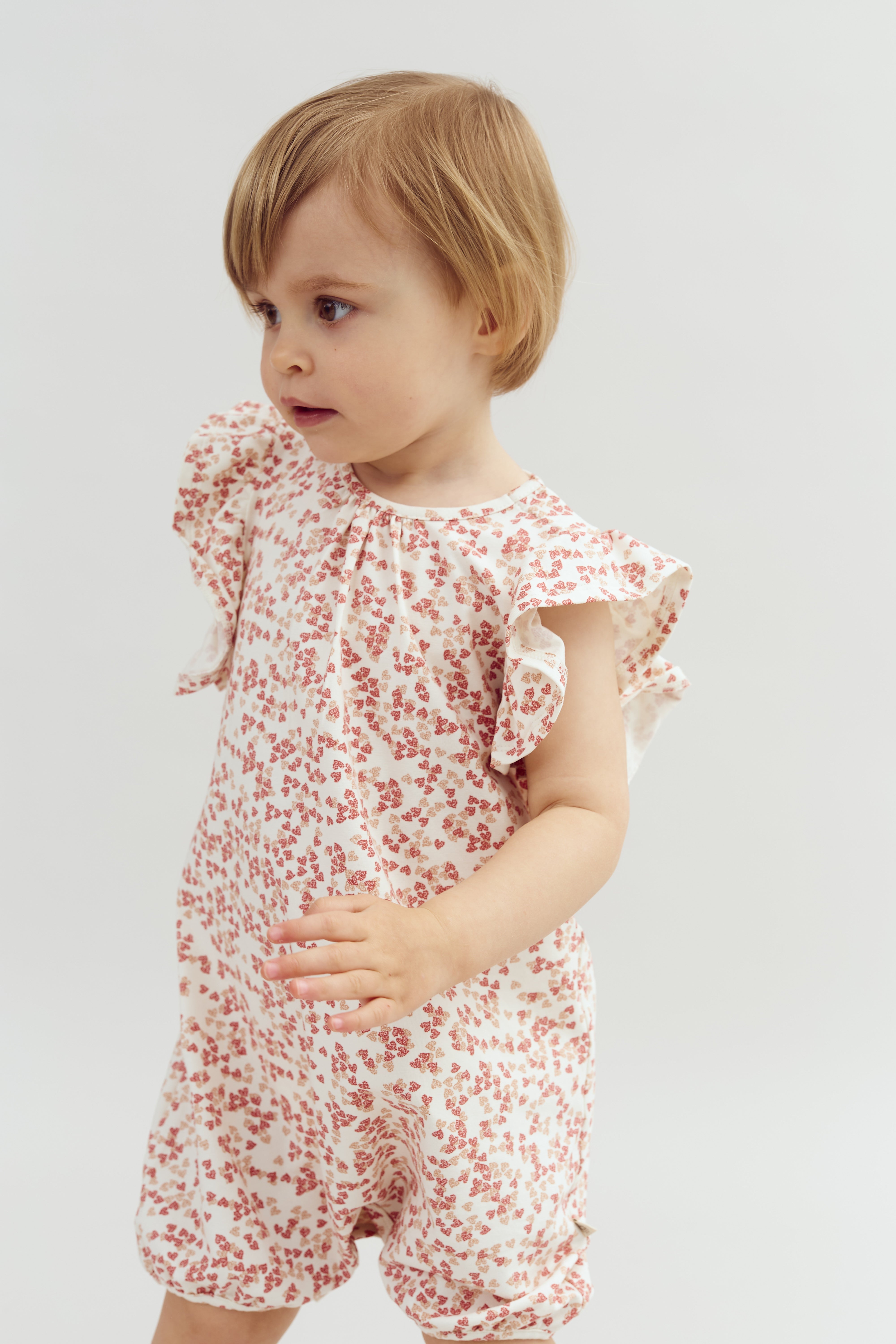 A child wearing a white outfit with red floral patterns looks to the side. The plain, light background highlights the child's expression and clothing details.