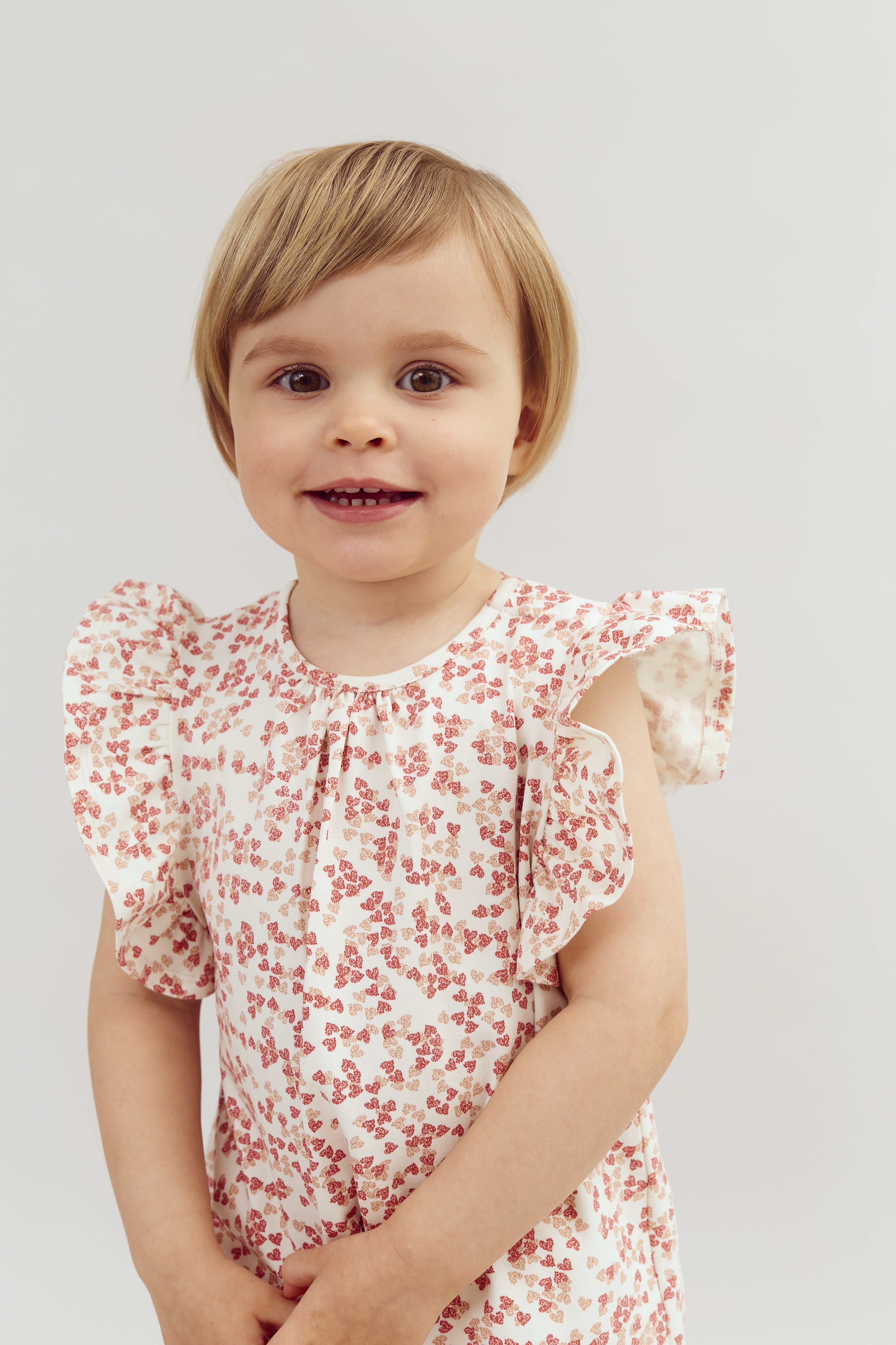 Child smiling while wearing a floral-patterned dress with ruffled sleeves, set against a plain, light background.