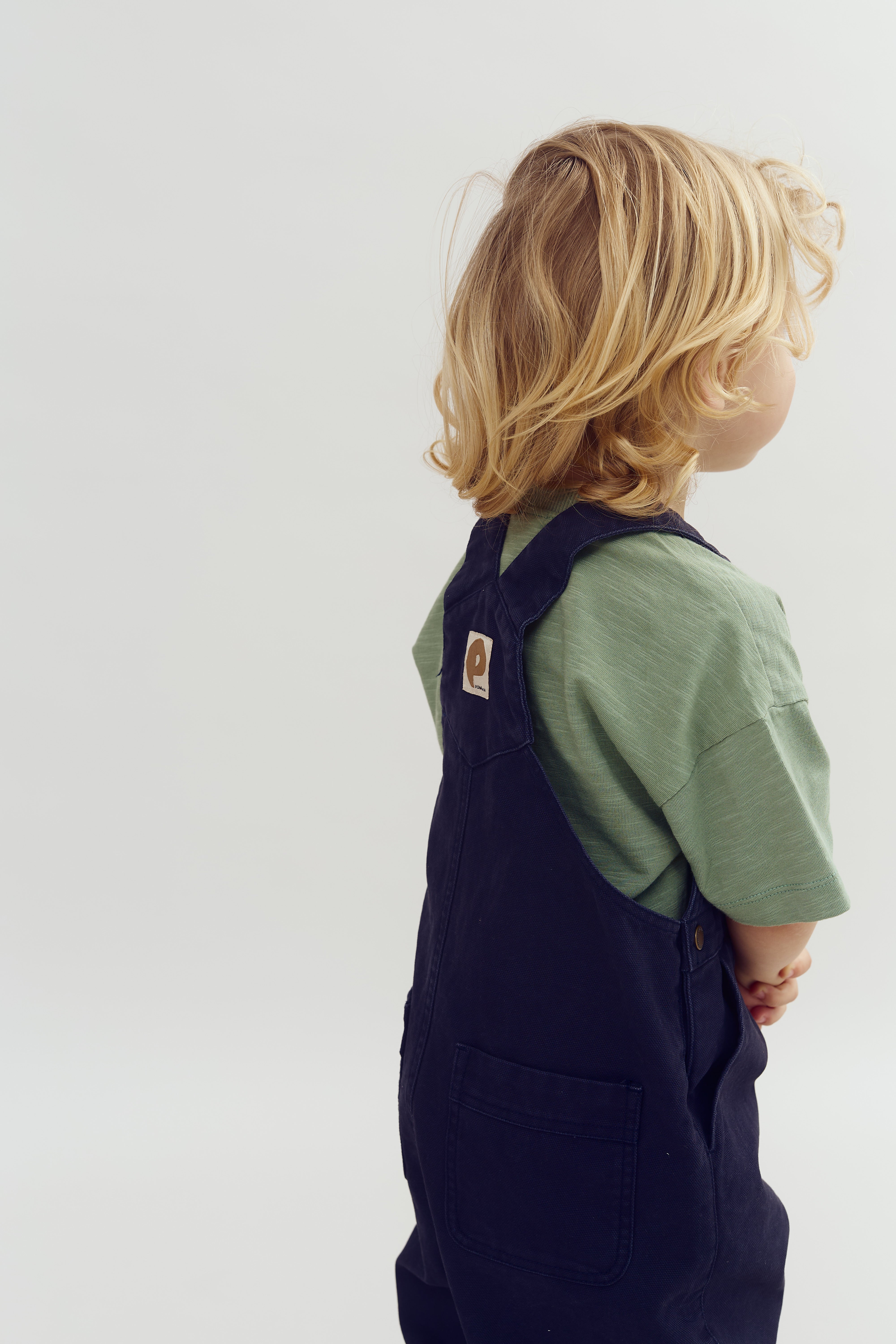 A child with light blond hair, wearing a green shirt and dark overalls, stands sideways with hands clasped, set against a plain, neutral background.