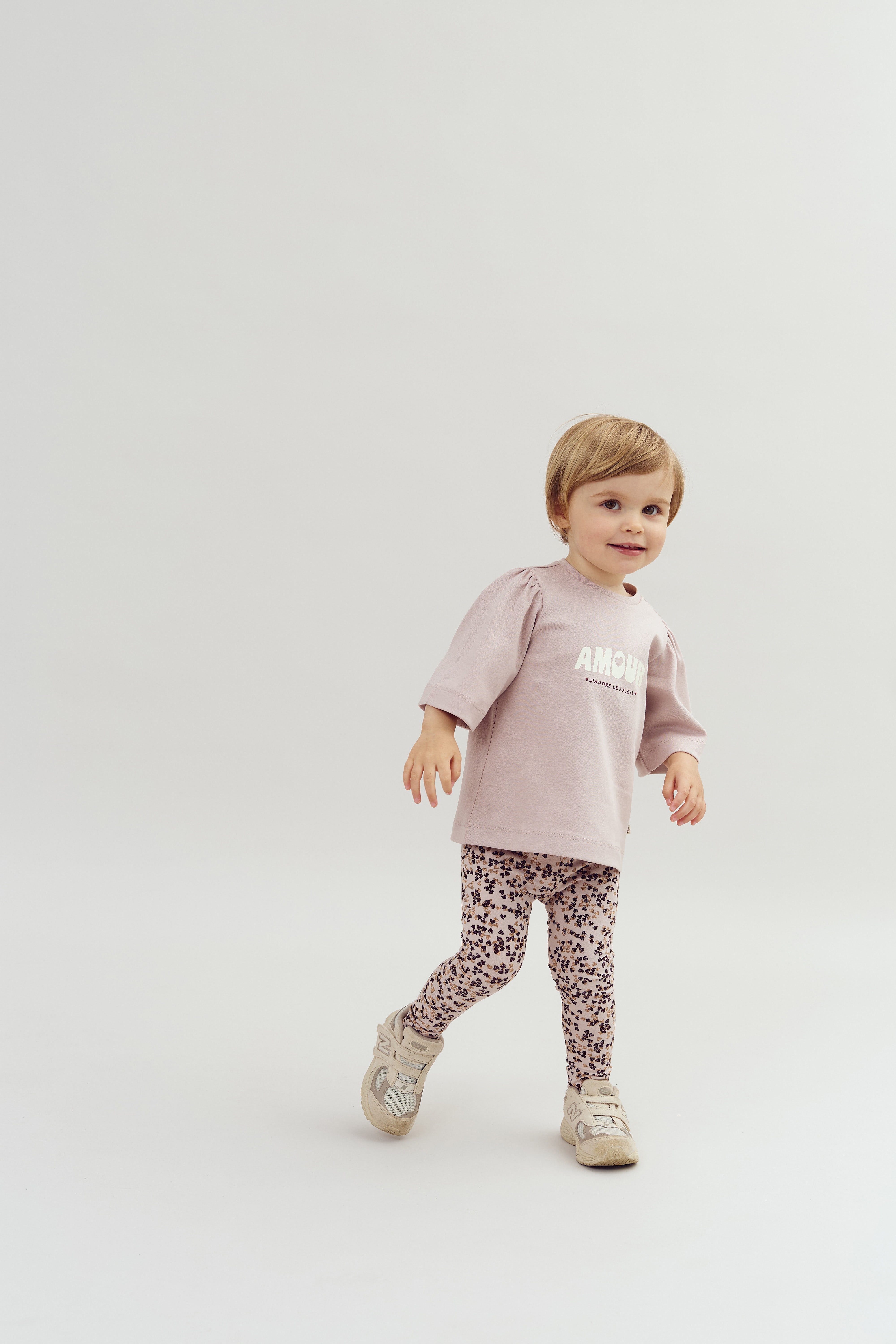 A young child walks confidently in a studio setting, wearing a pink top with the text "AMOUR" above floral leggings and light sneakers. The background is plain white.