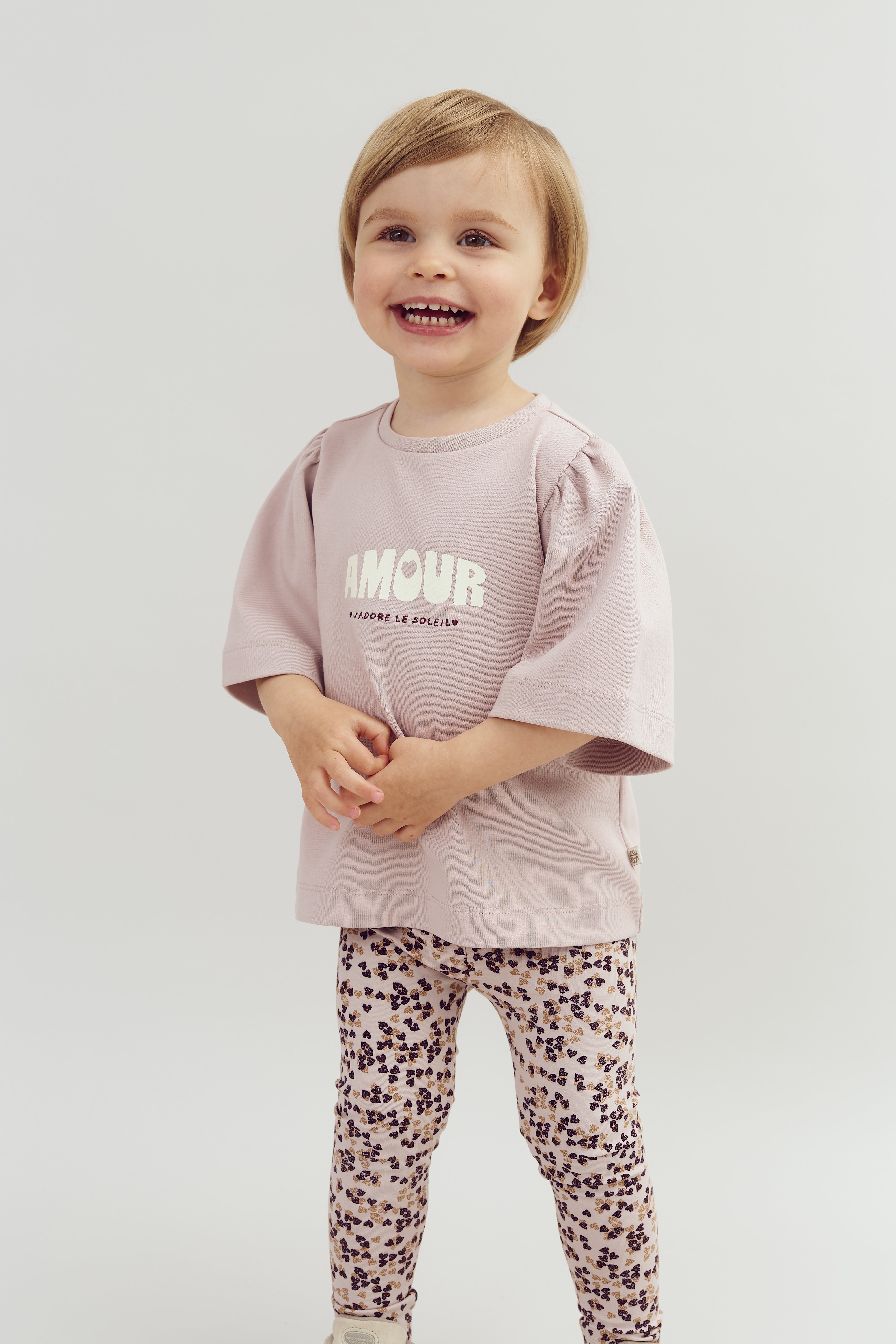 A young child is smiling, wearing a light pink shirt that says "AMOUR L'ADORE LE SOLEIL" and floral-patterned leggings, standing against a plain white background.