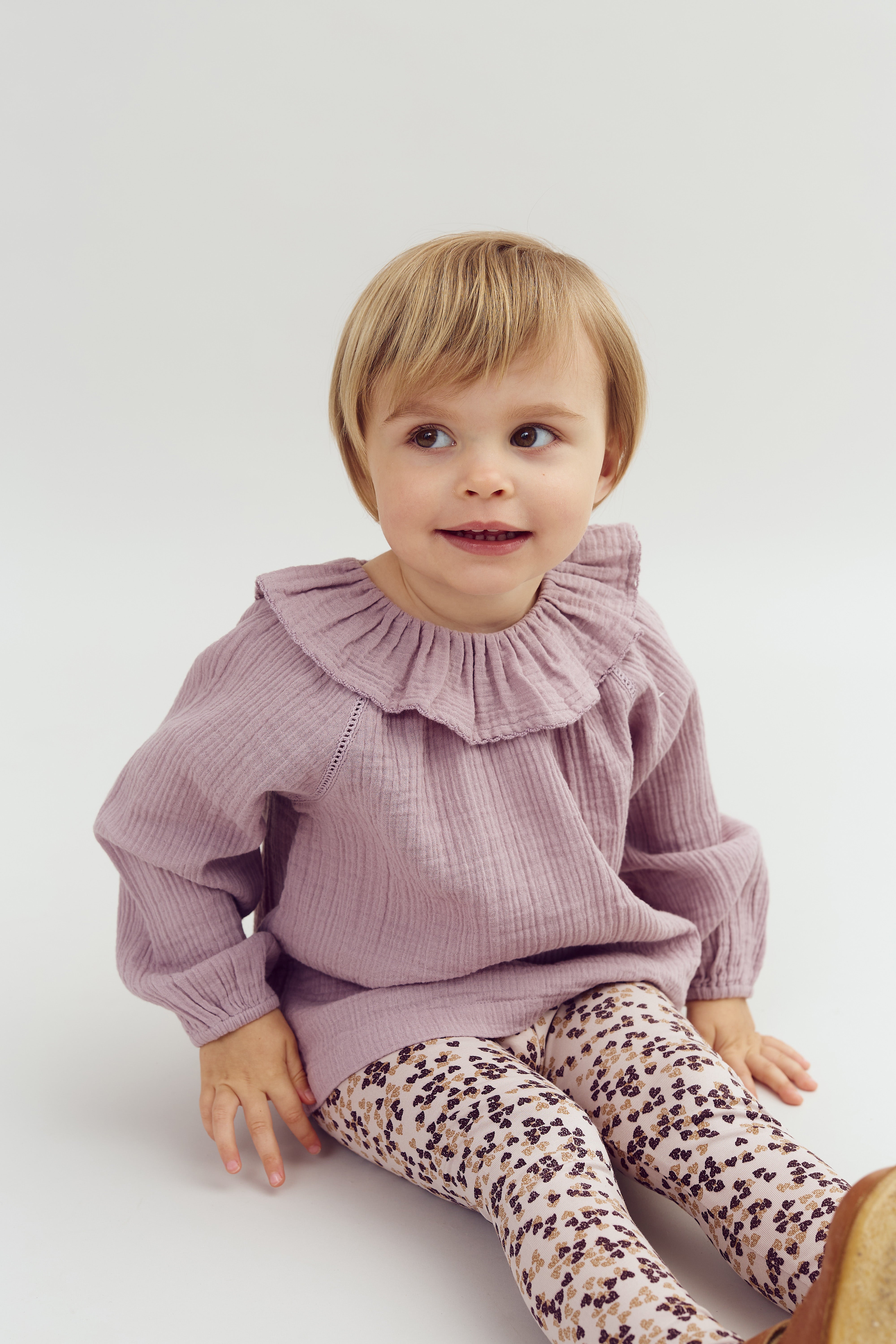A child sits and smiles, wearing a lilac sweater with a ruffled collar and floral-patterned leggings, in a neutral, well-lit setting.